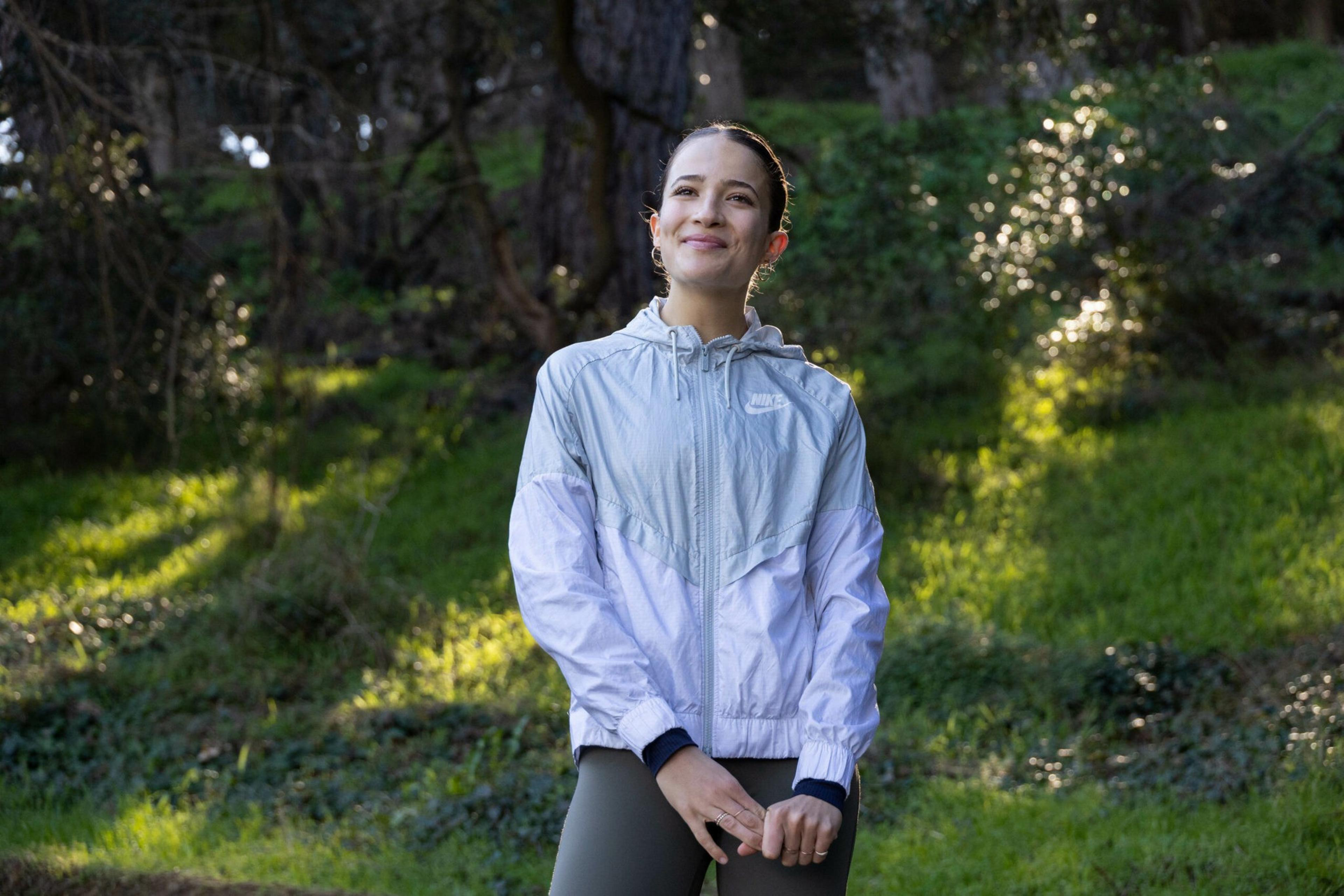 A woman stands outdoors in a light gray Nike jacket and dark leggings, smiling gently with trees and sunlight filtering through in the background.
