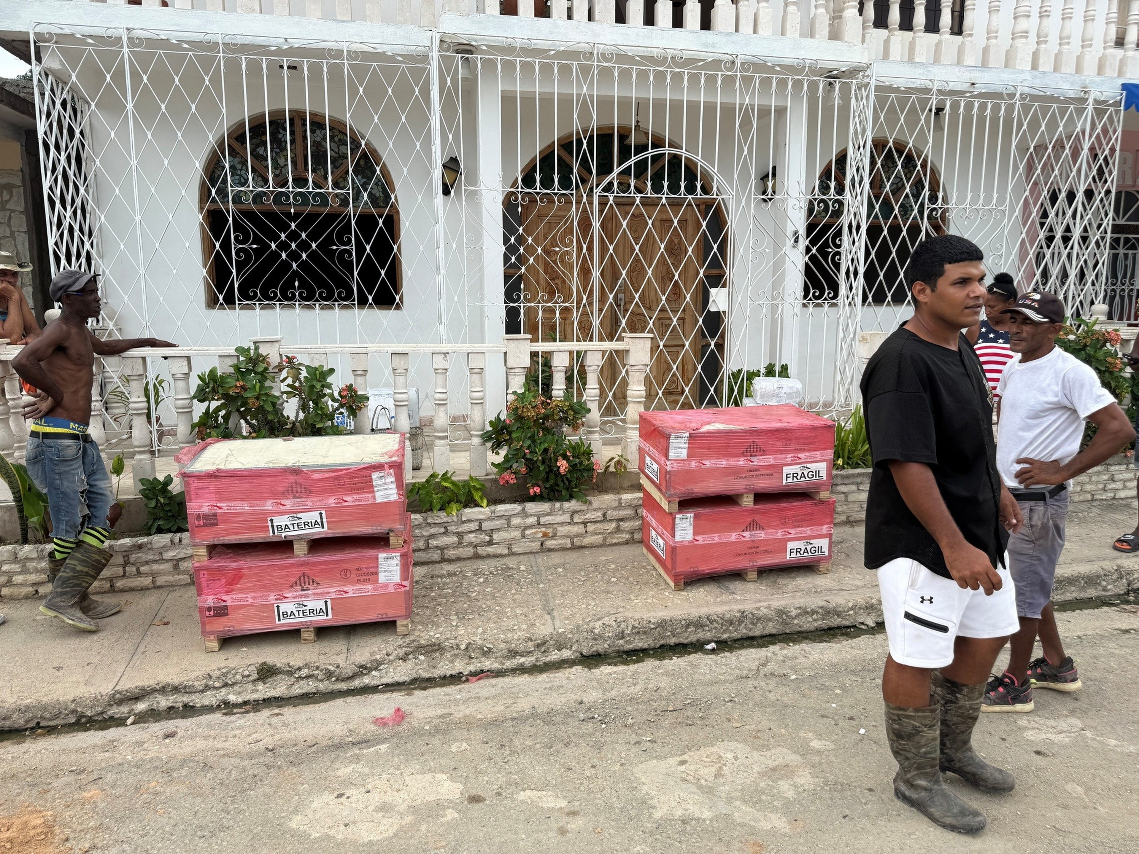 Four men stand and talk near four large, pink-wrapped boxes labeled “bateria” and “fragil” outside a white building with barred windows and plants.