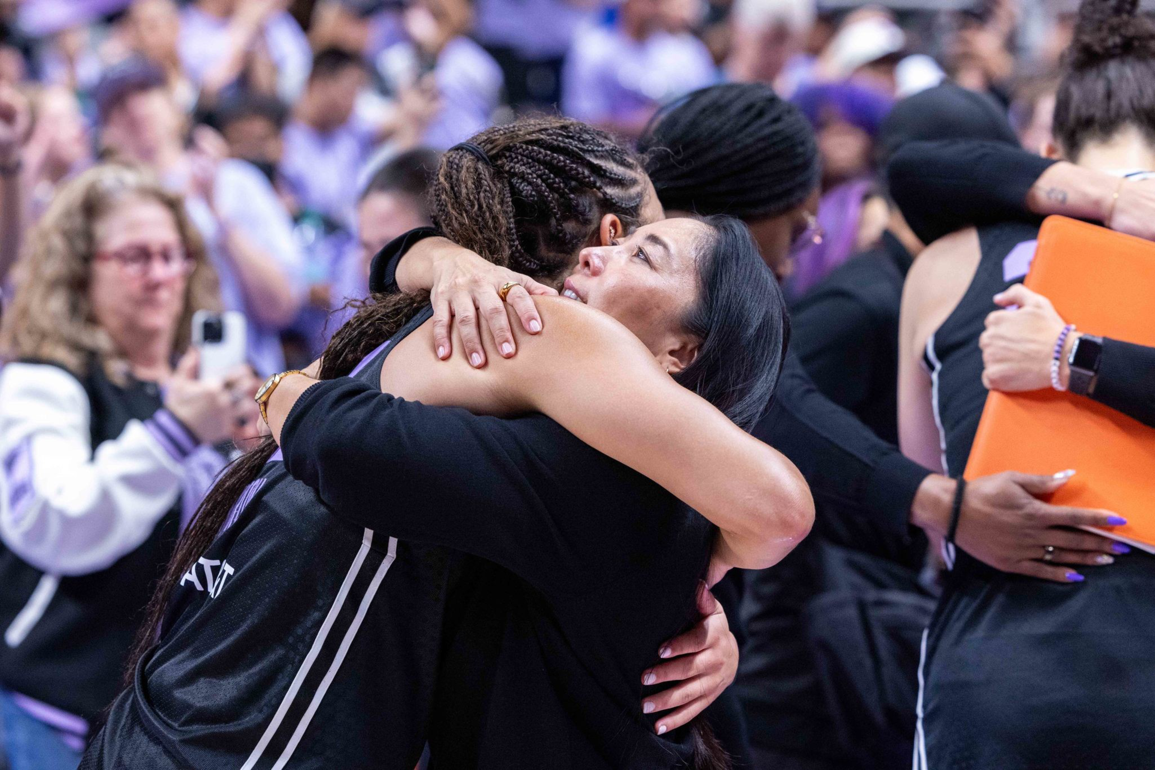 Two women embrace tightly in a crowded setting, with people in the background also hugging and showing emotional support.