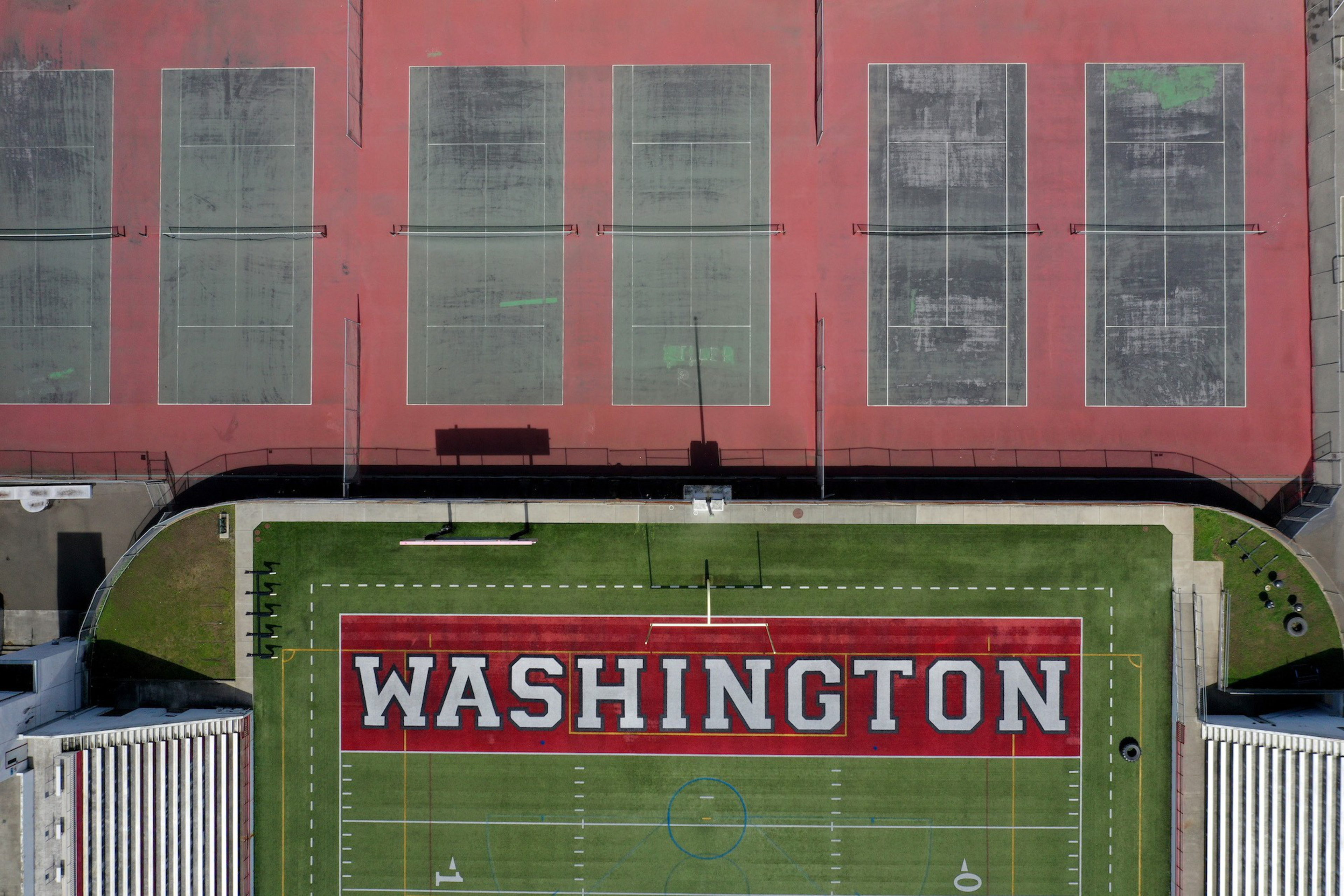 Aerial view of a sports complex with a green field marked "WASHINGTON" in white on a red background and six red tennis courts above it.