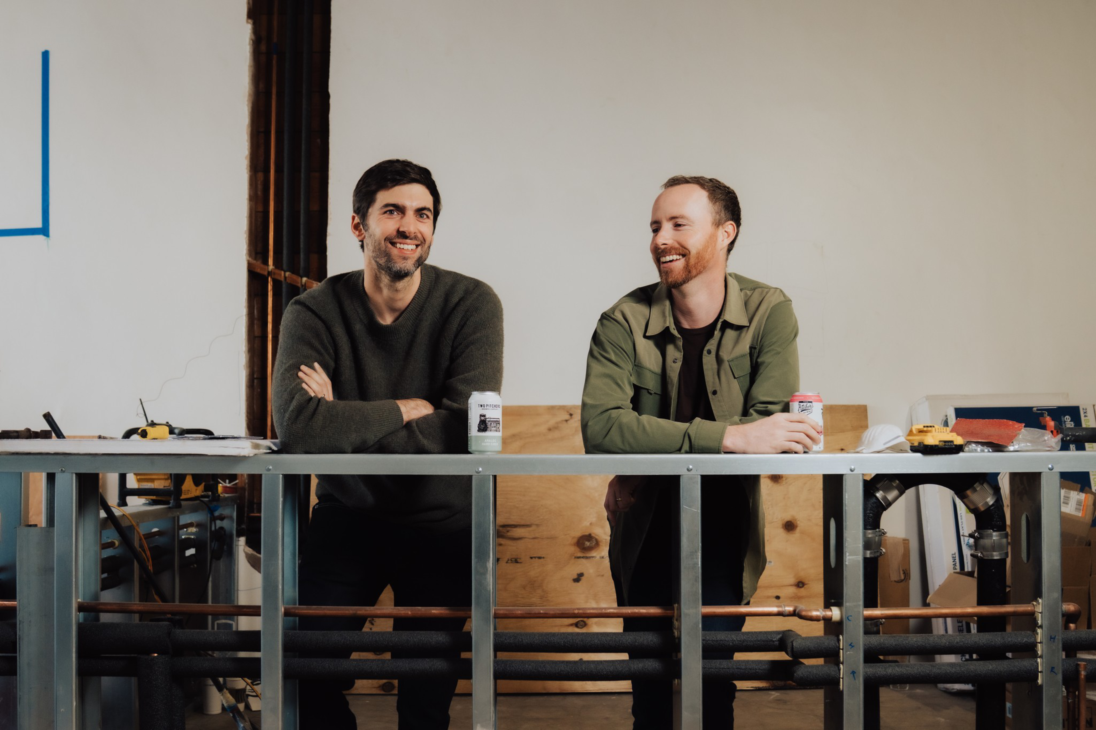 Two men lean on a metal railing inside a room, each holding a canned drink, smiling and looking at each other.
