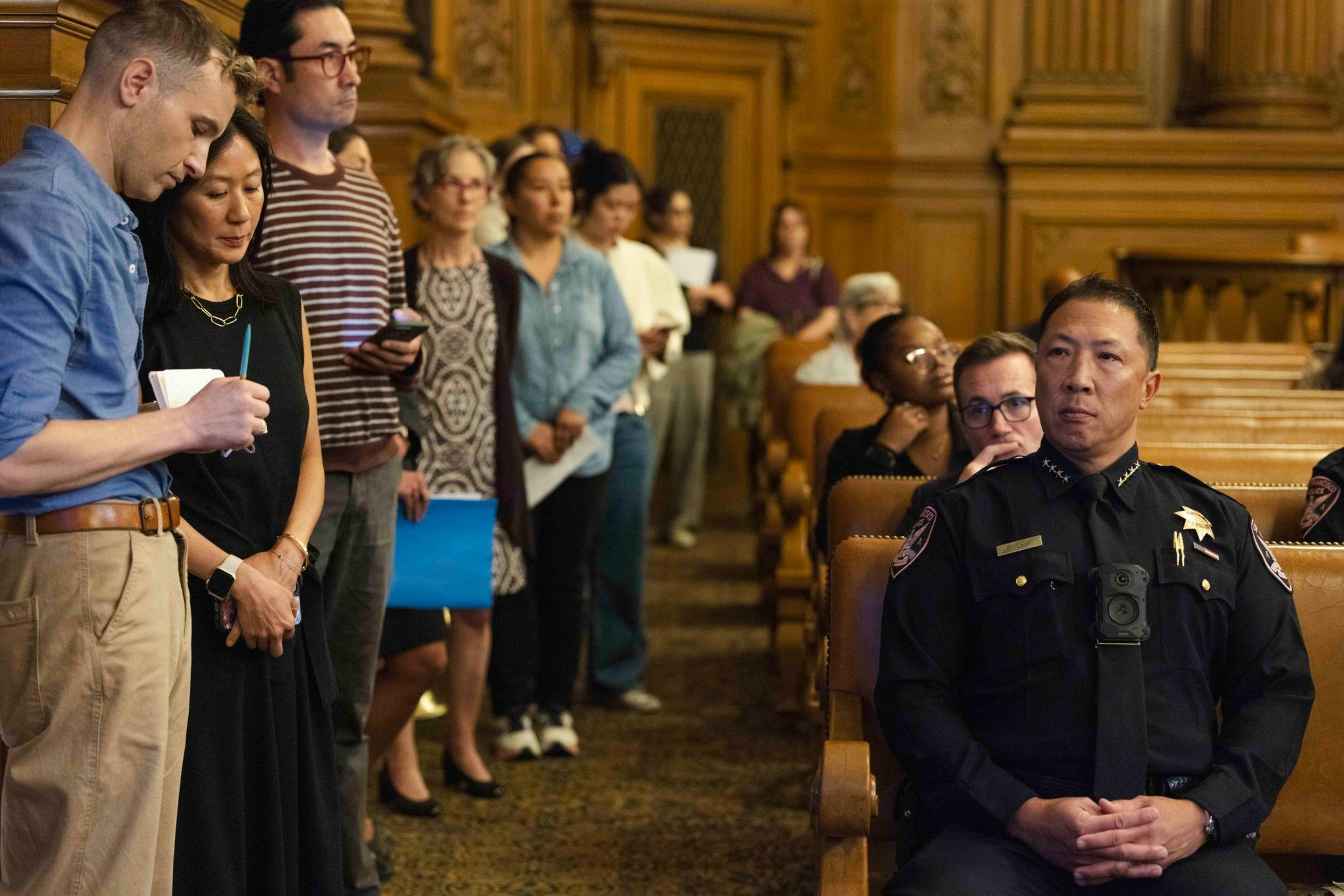 A group of people stand in line while a police officer in uniform sits in a wooden-paneled room with rows of benches.