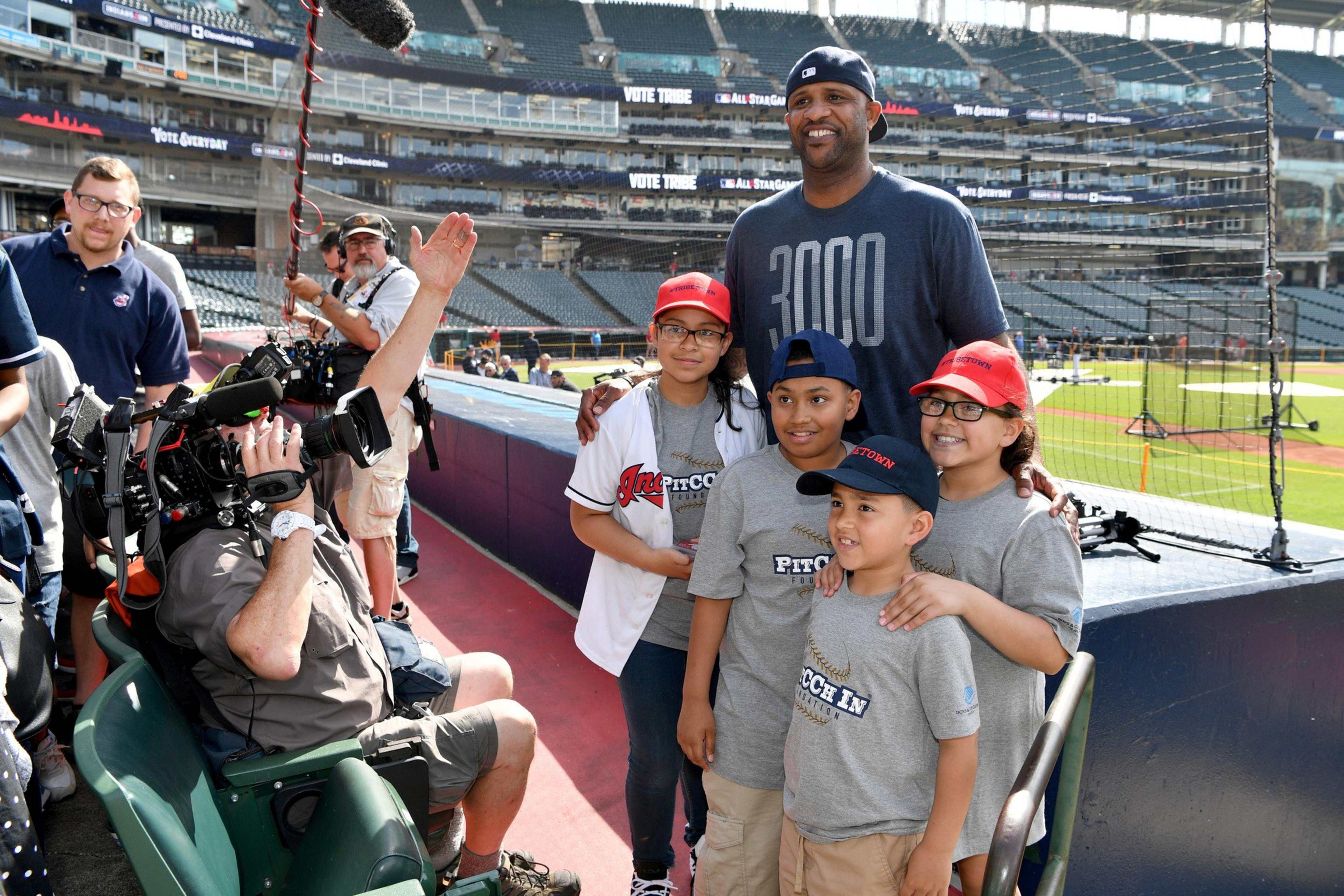 A man in a &quot;3000&quot; shirt stands with four smiling kids in baseball gear. They're posing for a photo near a baseball field, while a cameraman films nearby.
