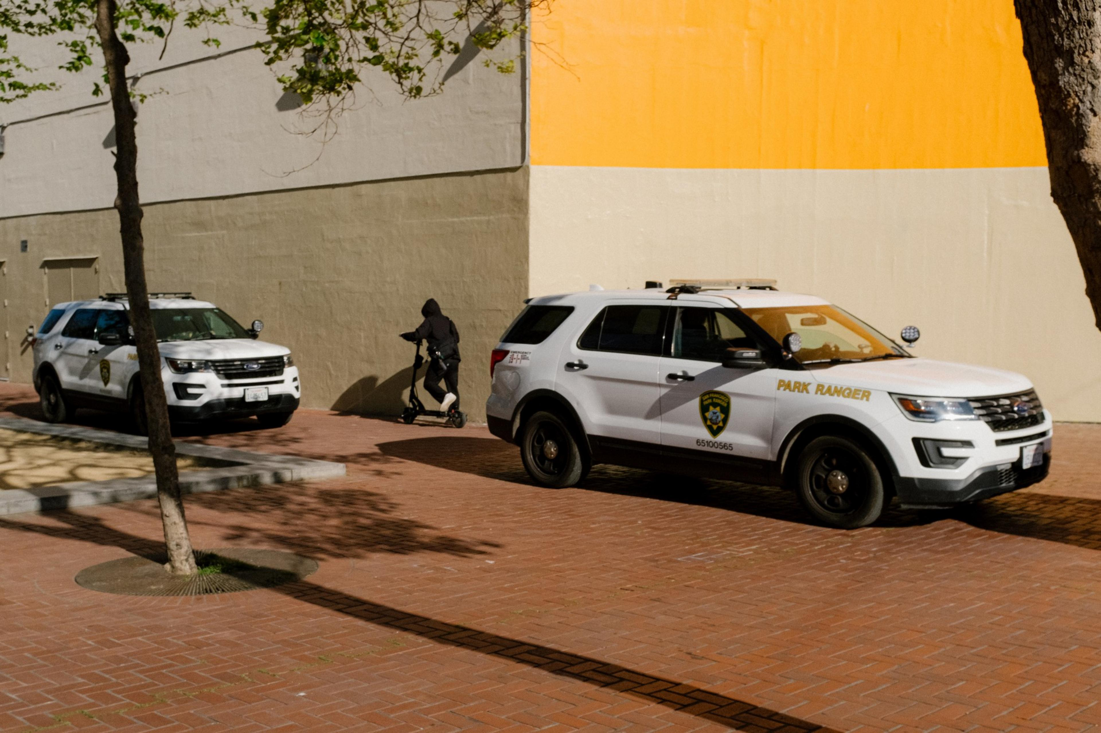 A person on a scooter passes by two parked park ranger vehicles near a large orange and white wall.