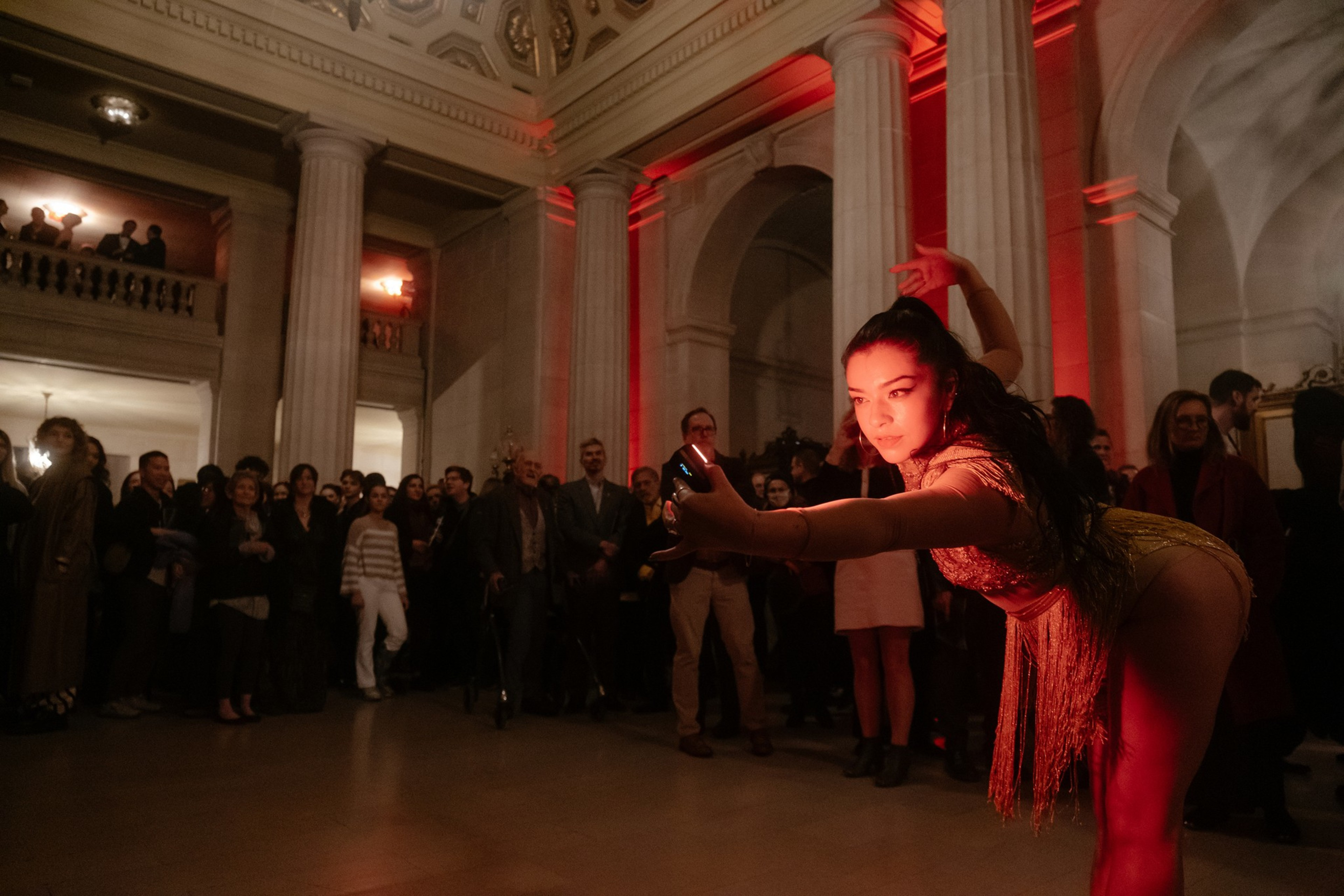 A dancer in a fringe outfit performs before an audience in a warmly lit grand hall.