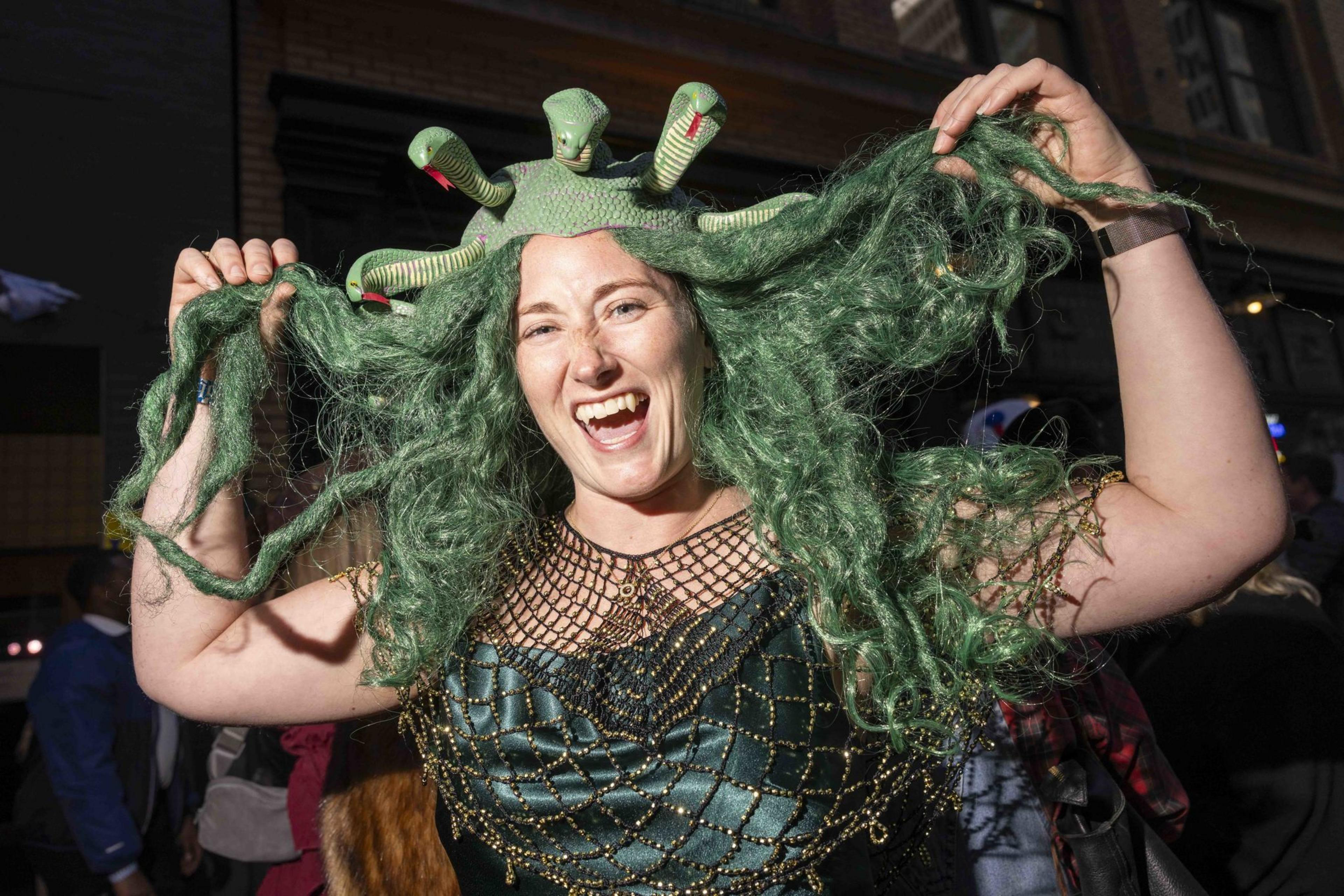 A person smiles widely while wearing a Medusa costume with green snake-like hair and a detailed green dress. They are holding their hair up playfully.