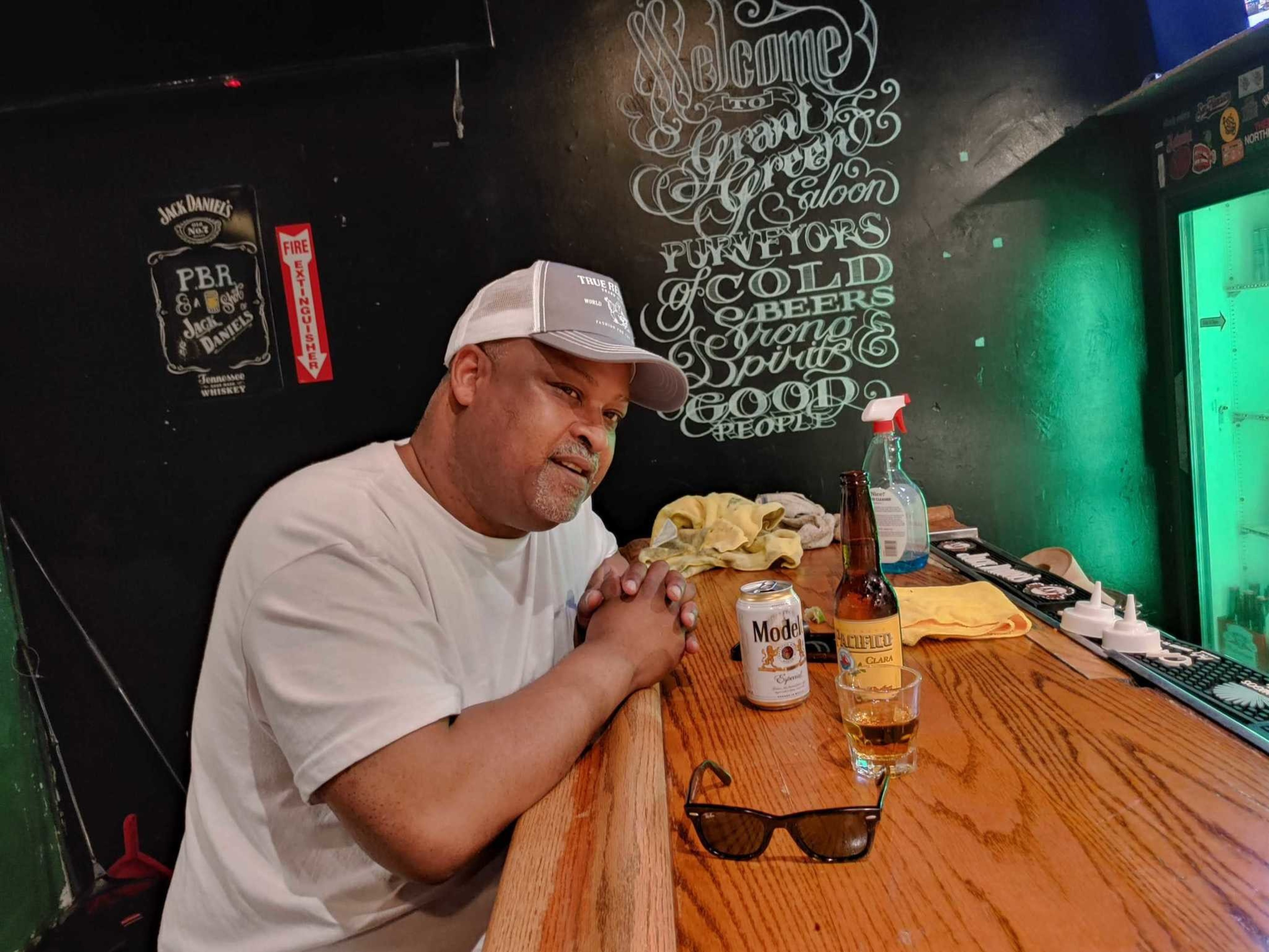 A man in a white shirt and cap sits at a wooden bar with a Modelo beer can, a Pacifico beer bottle, a shot glass, and sunglasses on the counter.