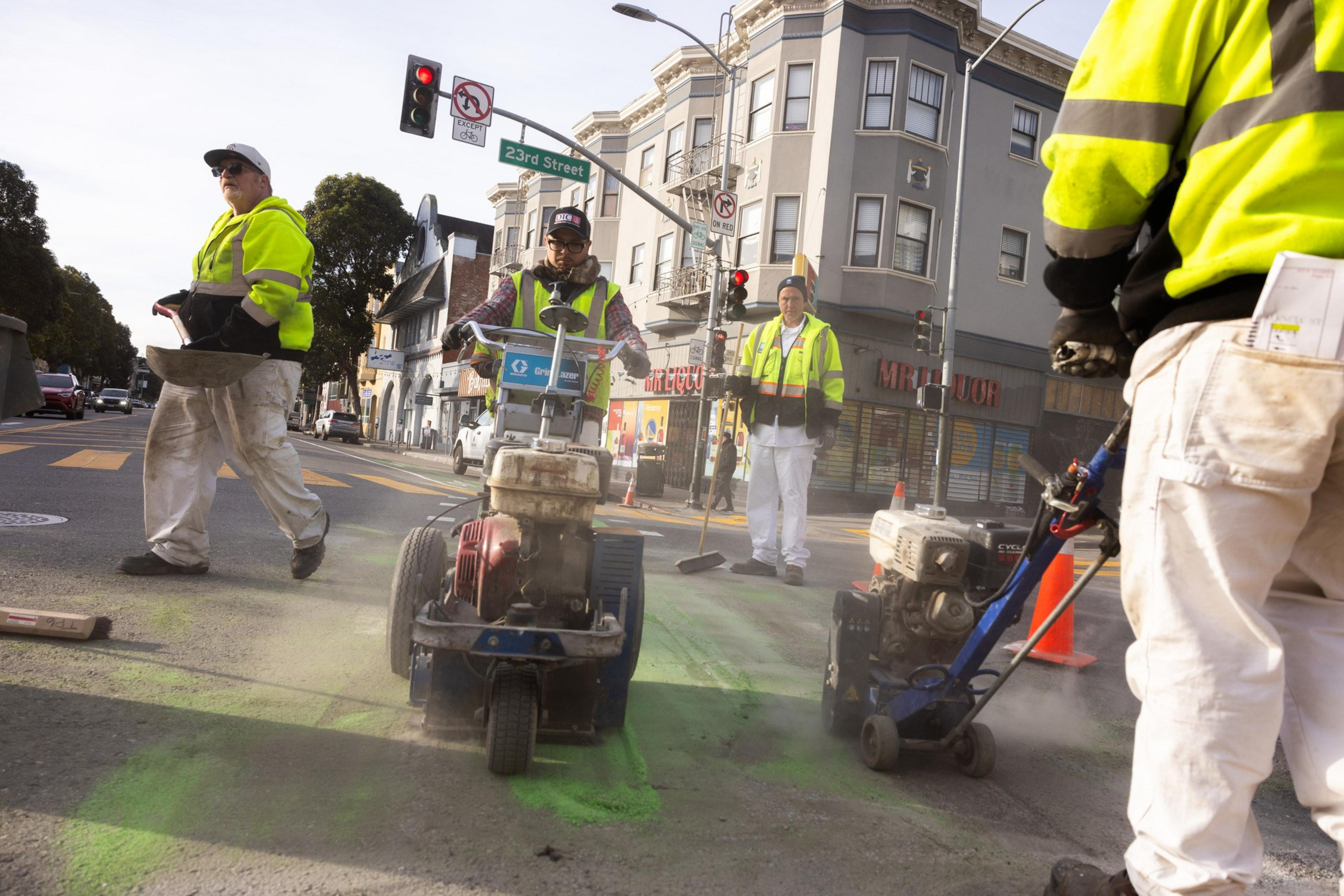 Workers in yellow vests use machines to paint green markings on a city street. They are near an intersection with buildings and traffic lights.