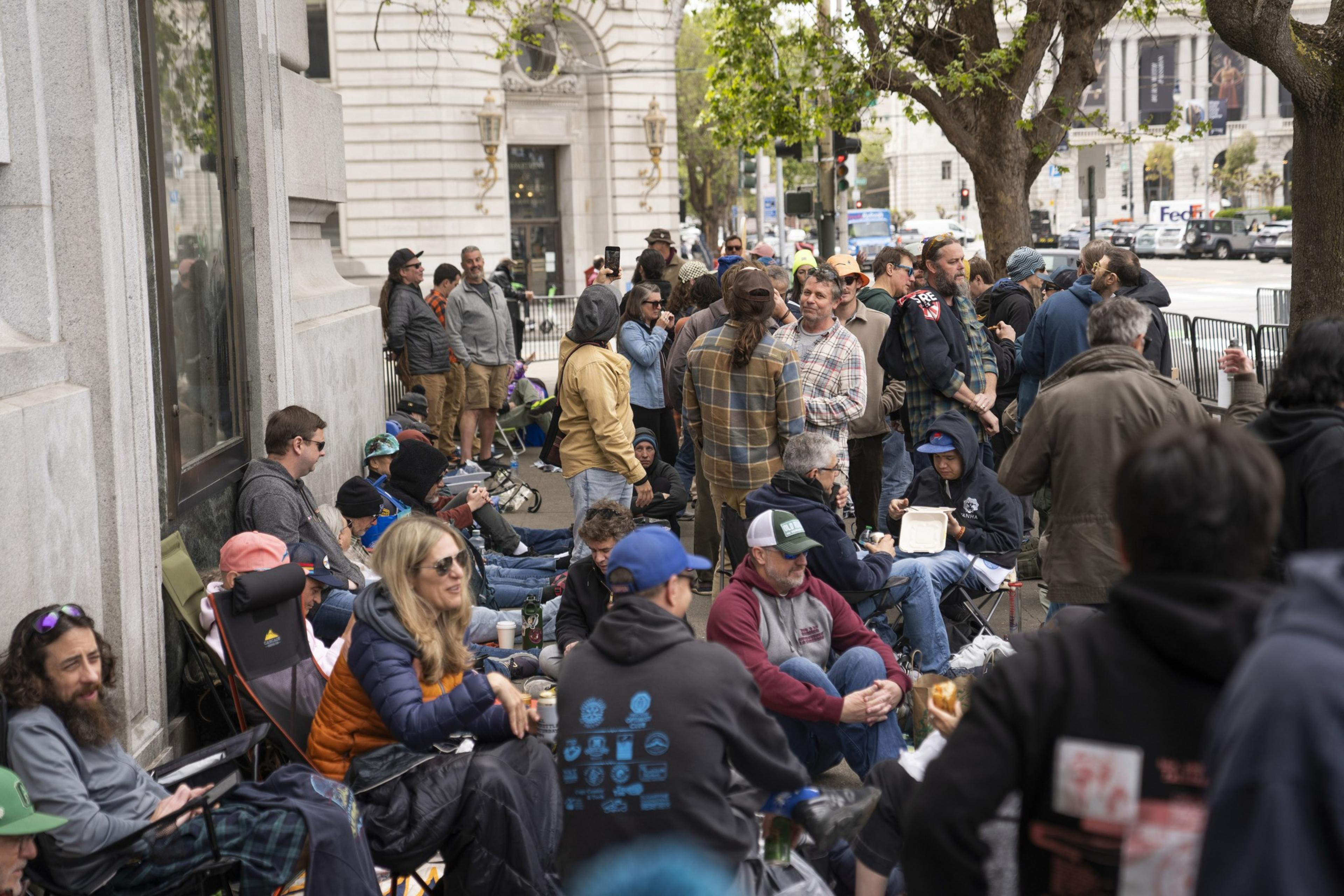 A large group of people is gathered outside a building. Some are seated in portable chairs, while others stand in small groups, chatting. Trees line the street.