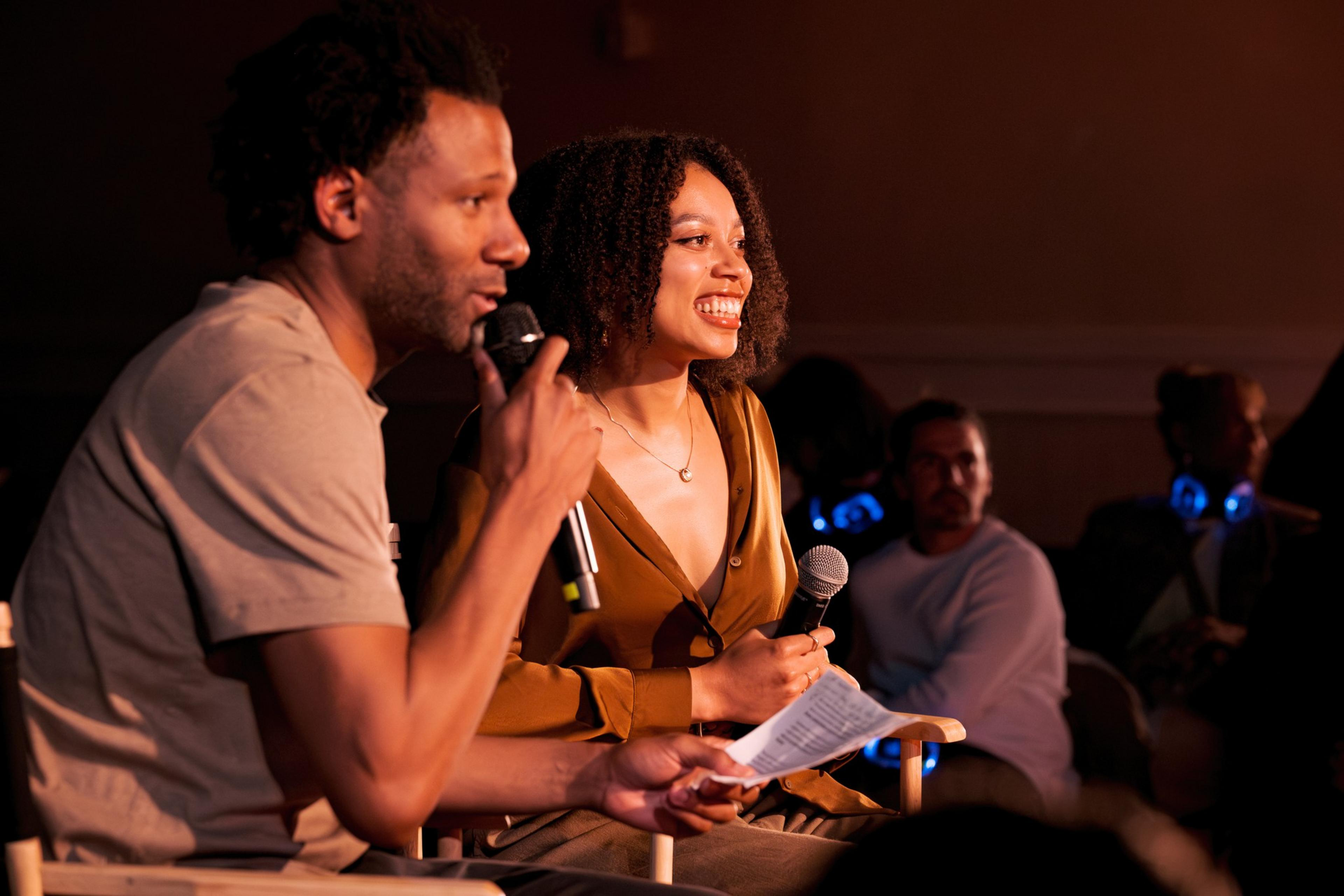 Two people are sitting side by side, holding microphones, with an audience in the background. They appear to be engaged in a lively conversation.