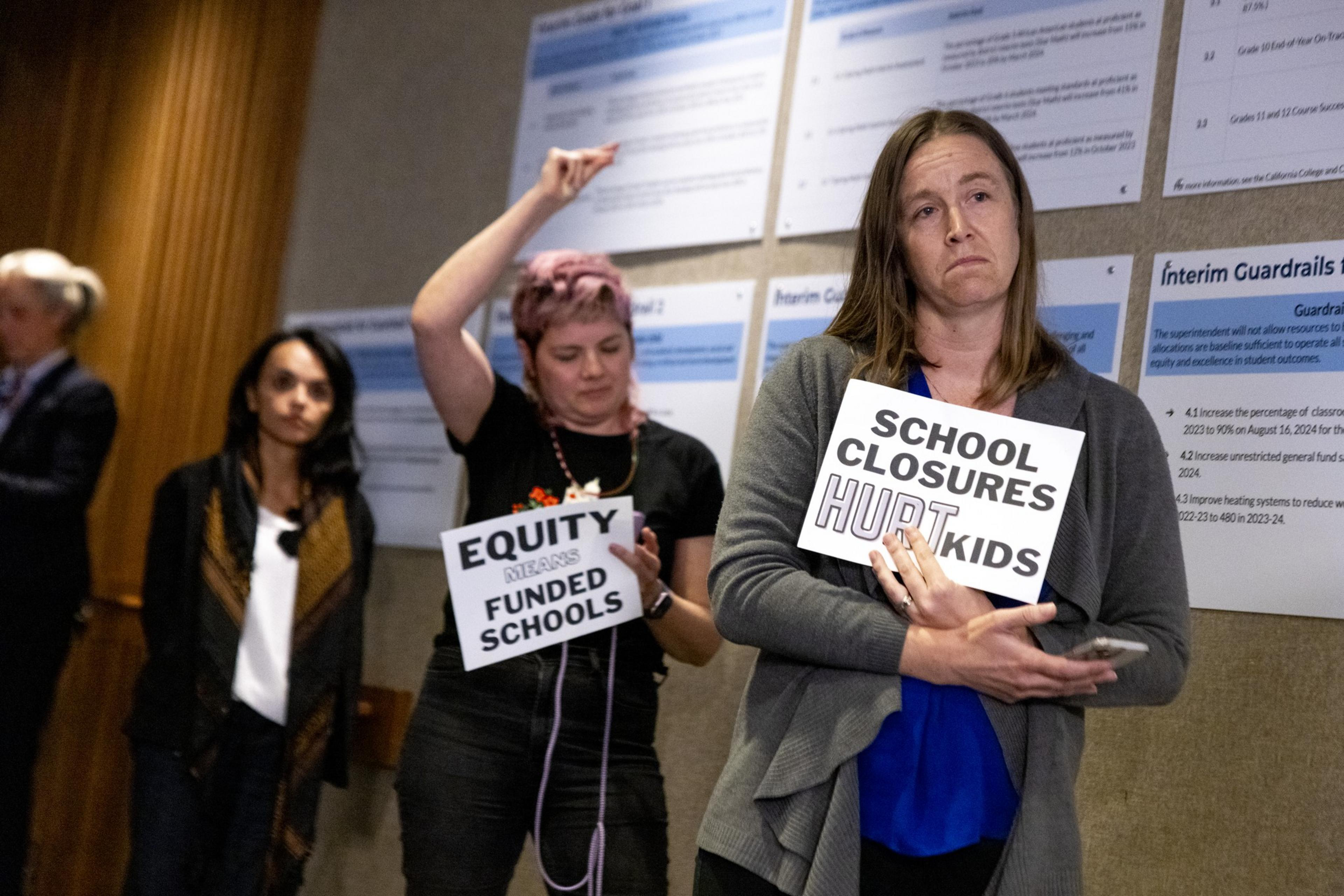 Three people hold signs at a meeting; one reads &quot;School closures hurt kids,&quot; and another reads &quot;Equity means funded schools.&quot; They appear concerned.