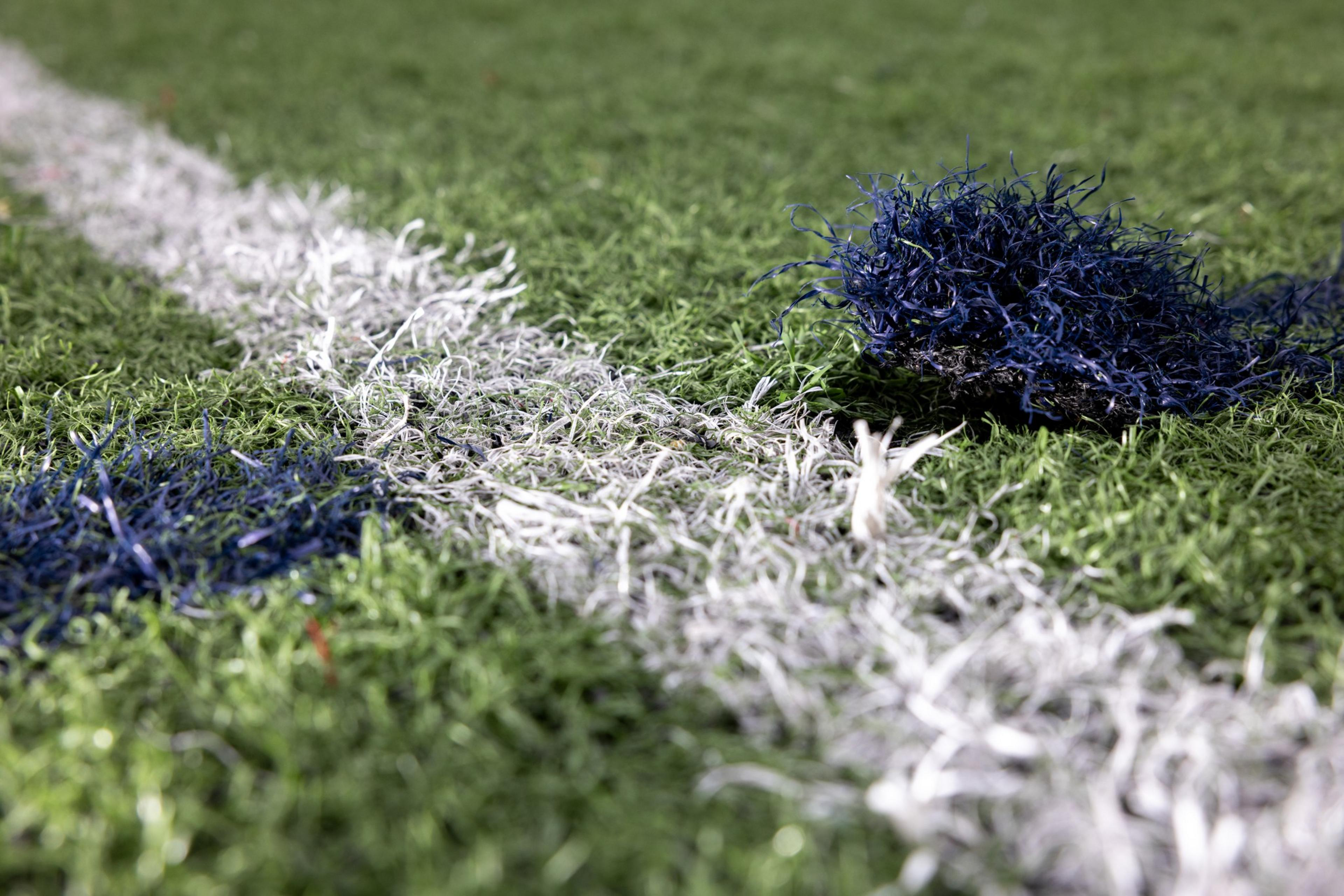 The image shows a close-up of artificial turf with green, blue, and white blades. A patch of blue blades is bunched up, suggesting possible damage.