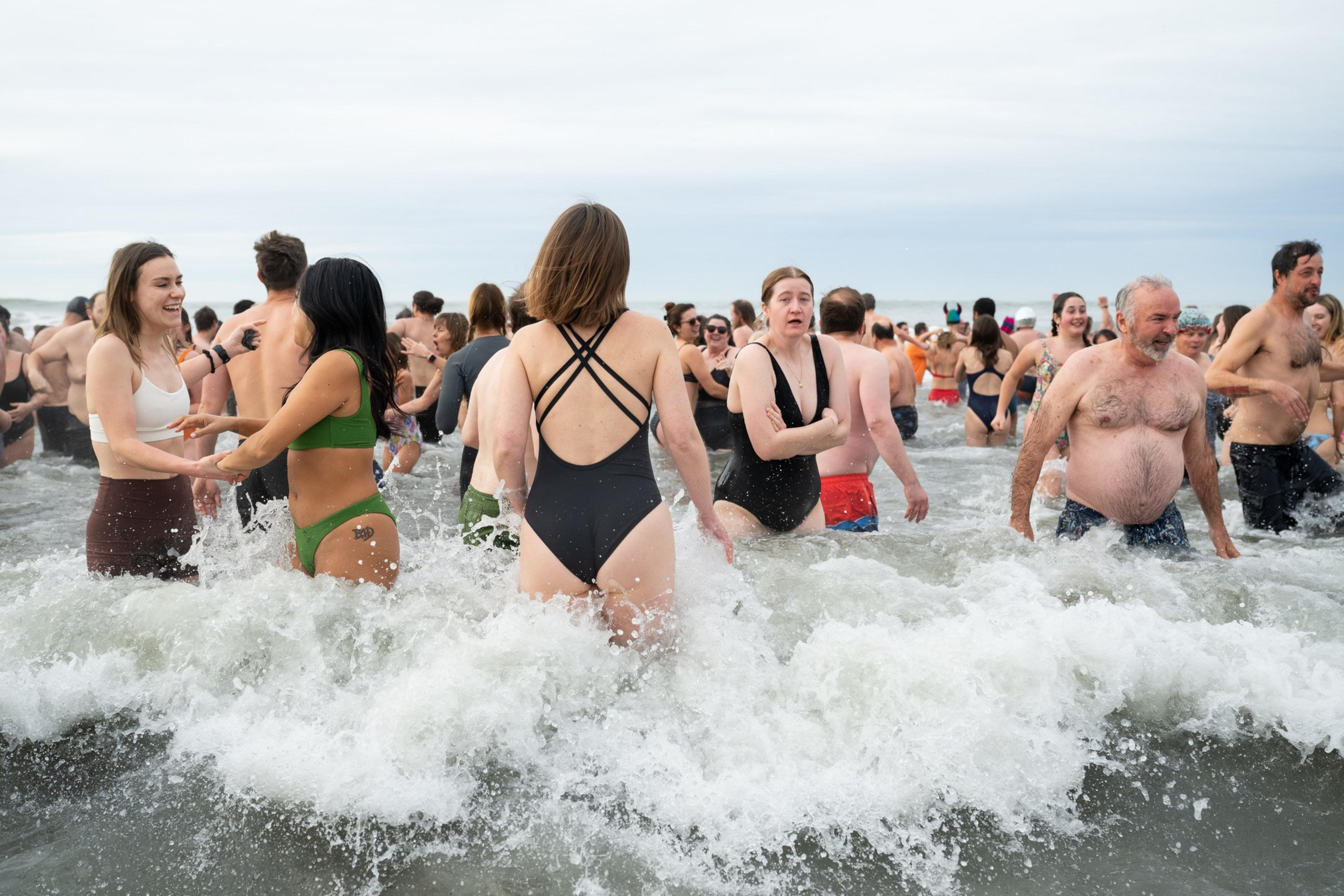 A group of people are joyfully wading in the ocean with waves splashing around them. They are dressed in swimsuits and appear to be enjoying a chilly swim.