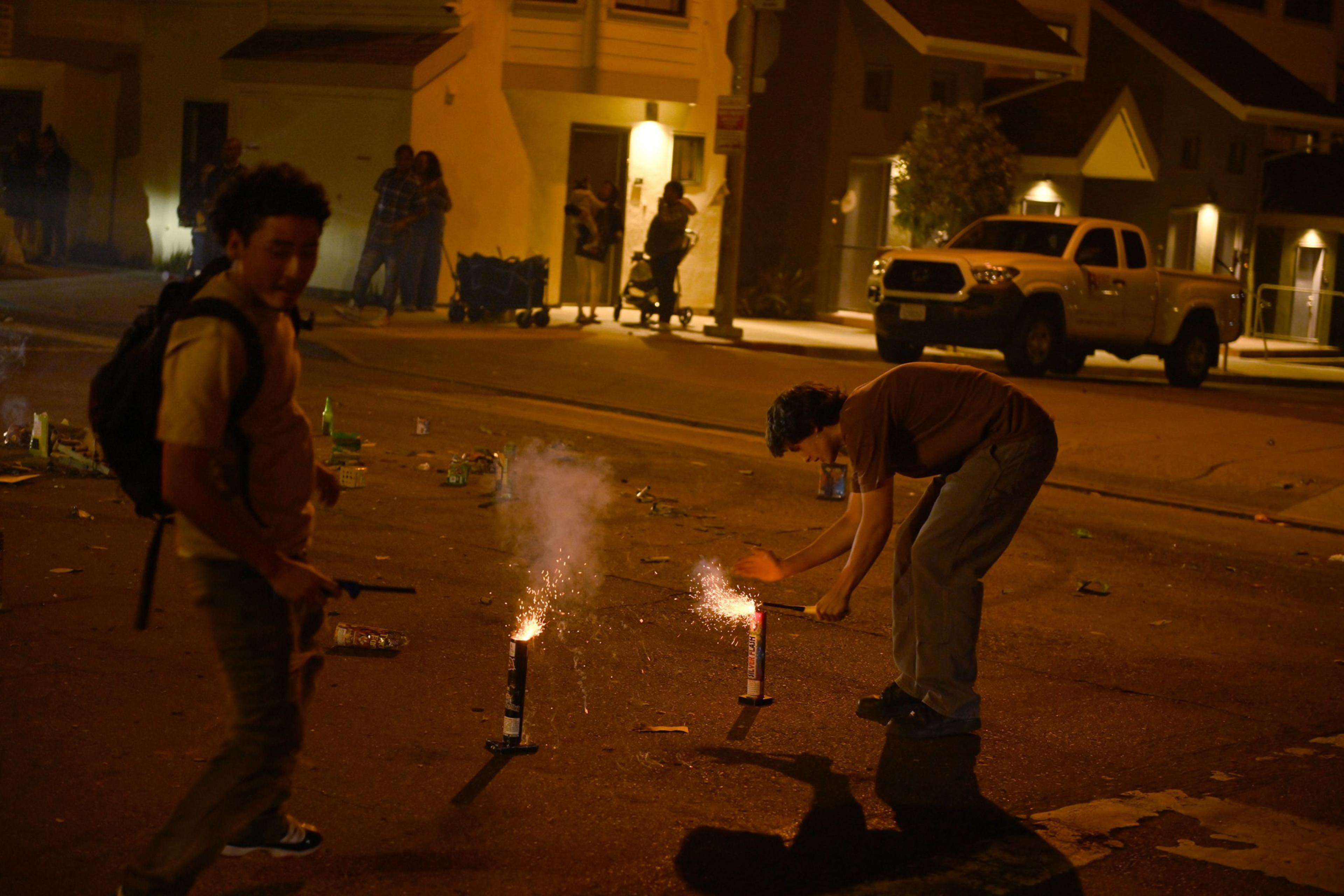 Several people gather at night, using smartphones to illuminate a man in a dark shirt and mask, who is holding a cylindrical object with a fuse.