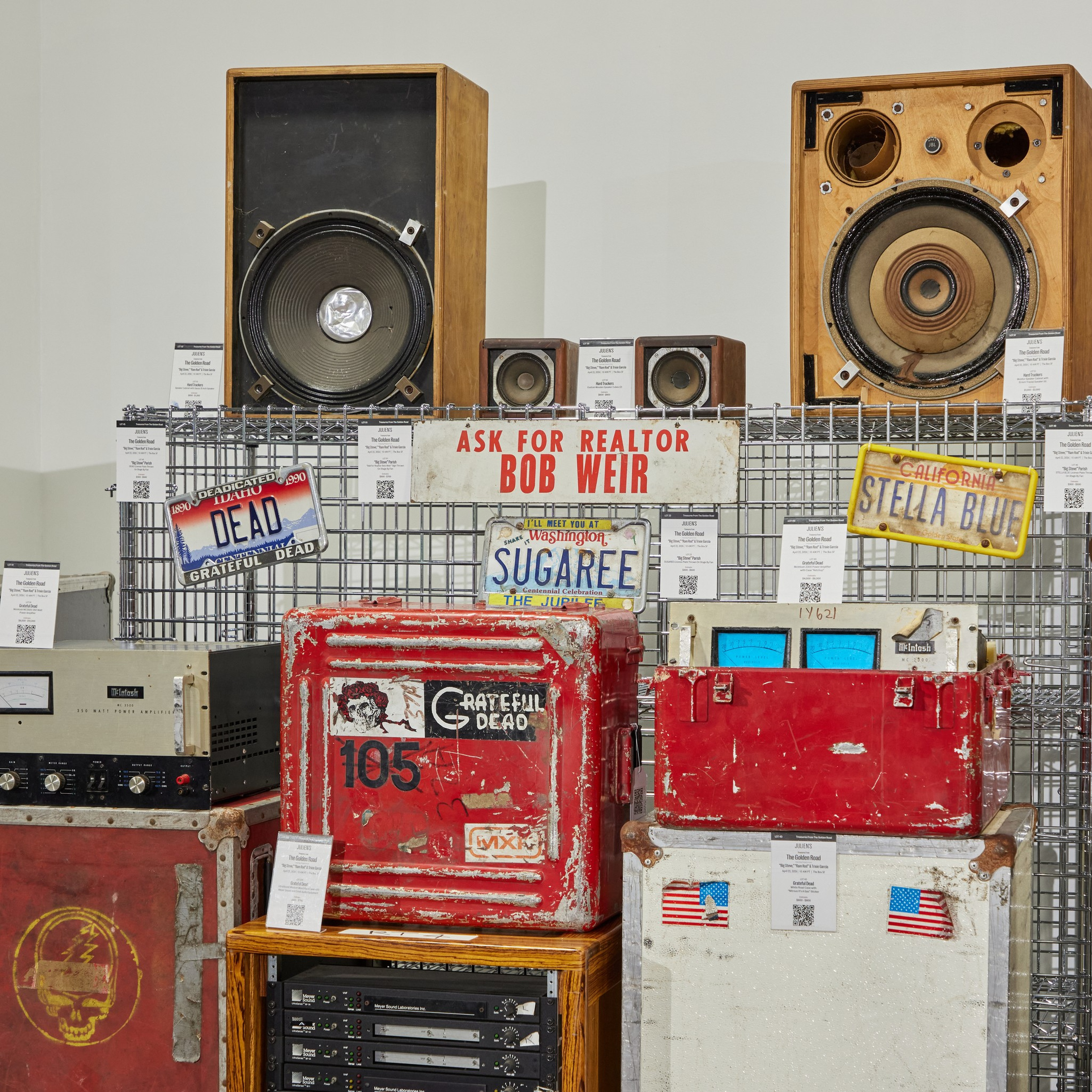 Vintage audio equipment including large wooden speakers, red and white crates with Grateful Dead logos, and several license plates and signs are displayed on a metal grid.
