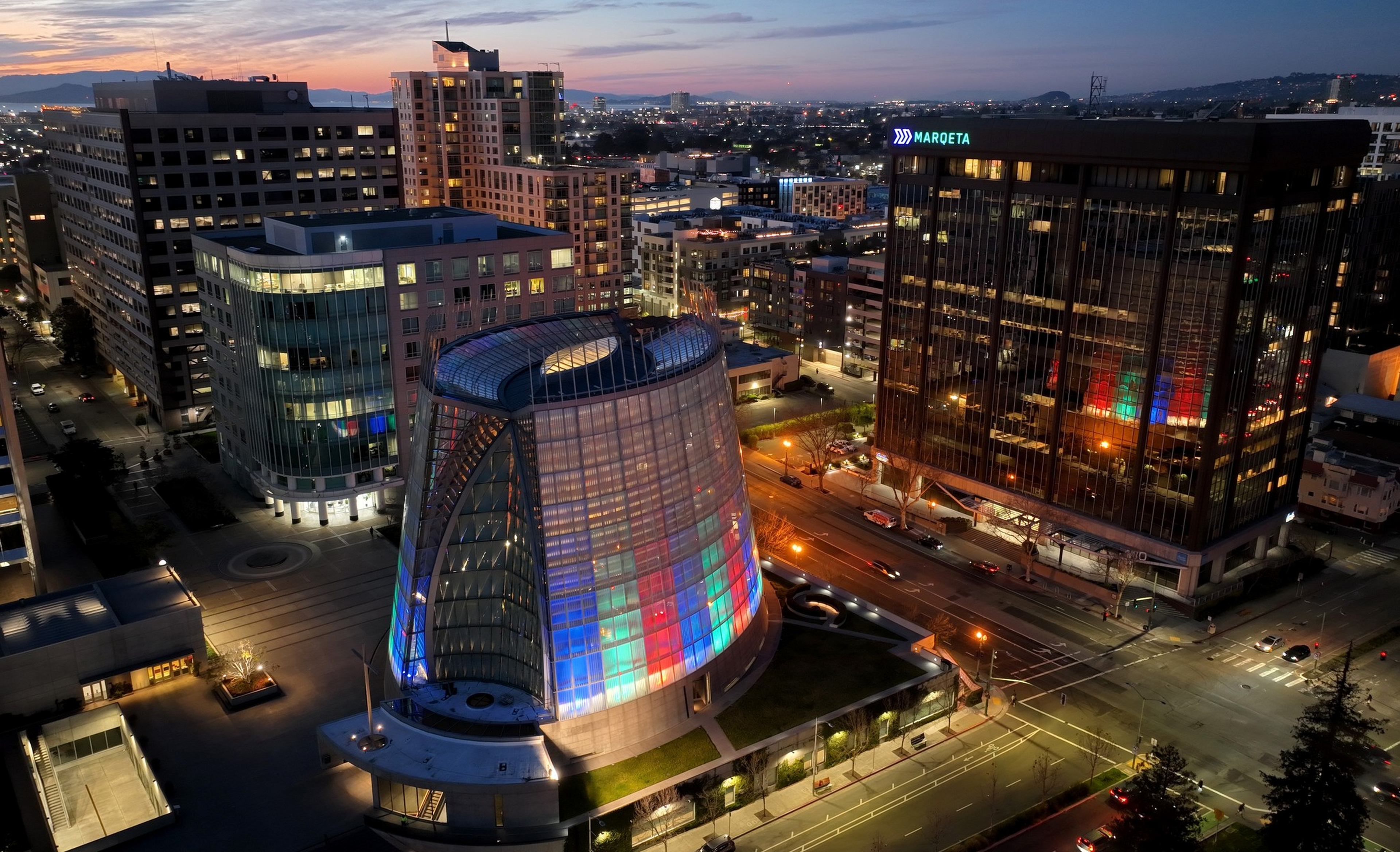 A modern cityscape at dusk features a unique curved glass building illuminated with colorful lights, surrounded by tall office buildings and streets.