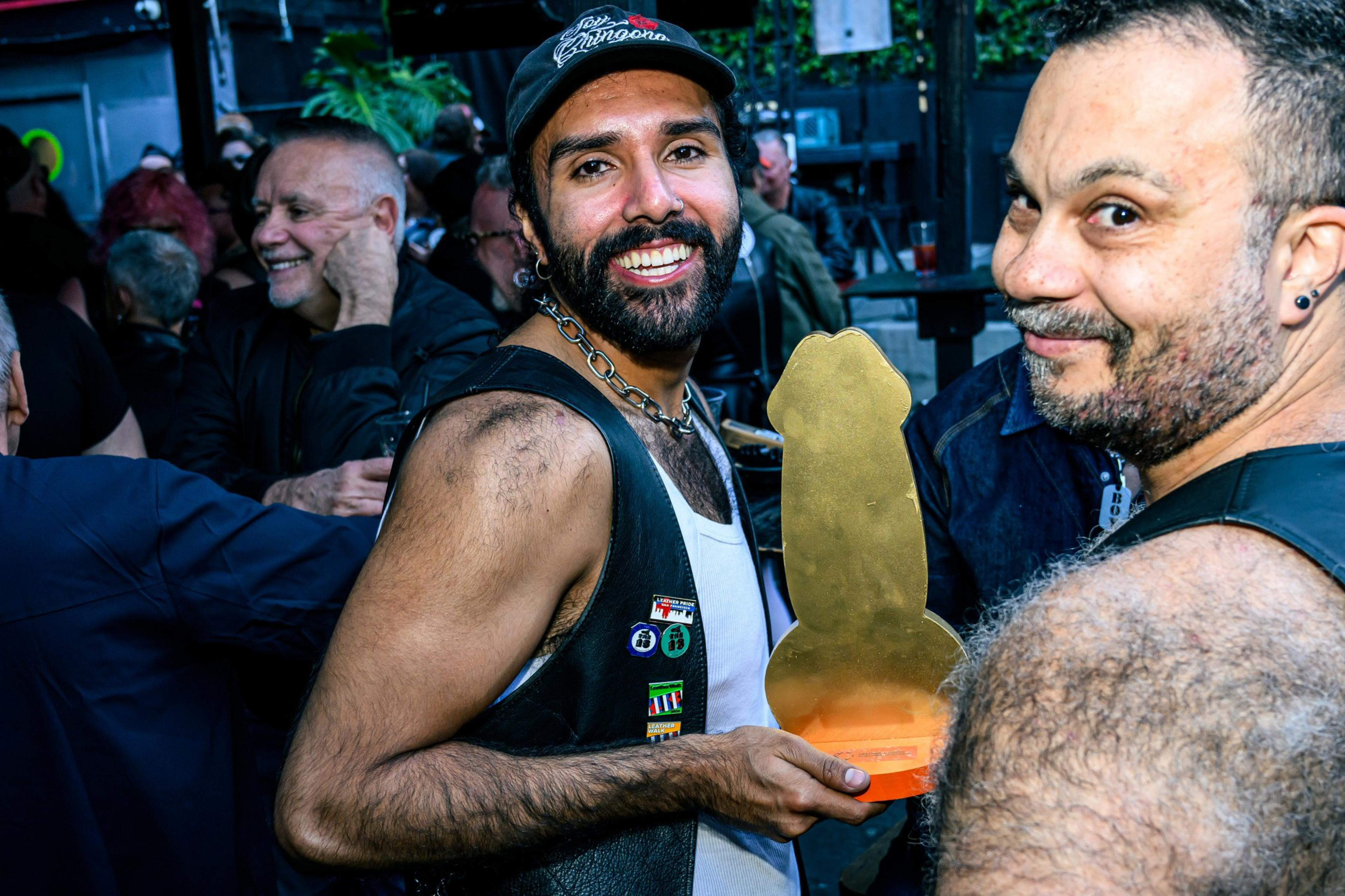 A man in a leather vest and hat smiles while holding a phallic-shaped trophy. Another man beside him, also smiling, is wearing a black top. People are in the background.