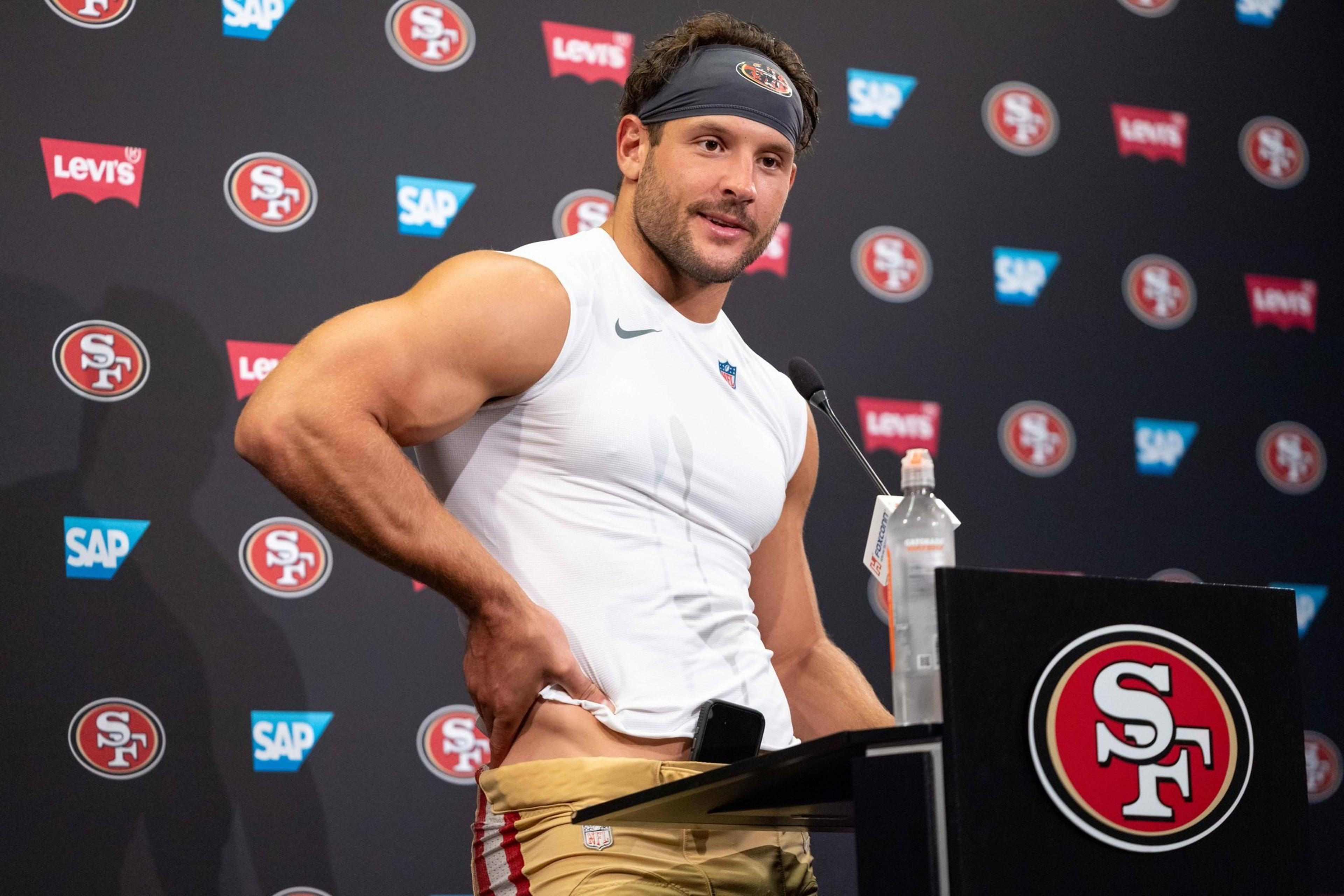 A muscular man in a sleeveless shirt and headband stands at a podium with an SF logo, adjusting his waistband. A branded backdrop is behind him.