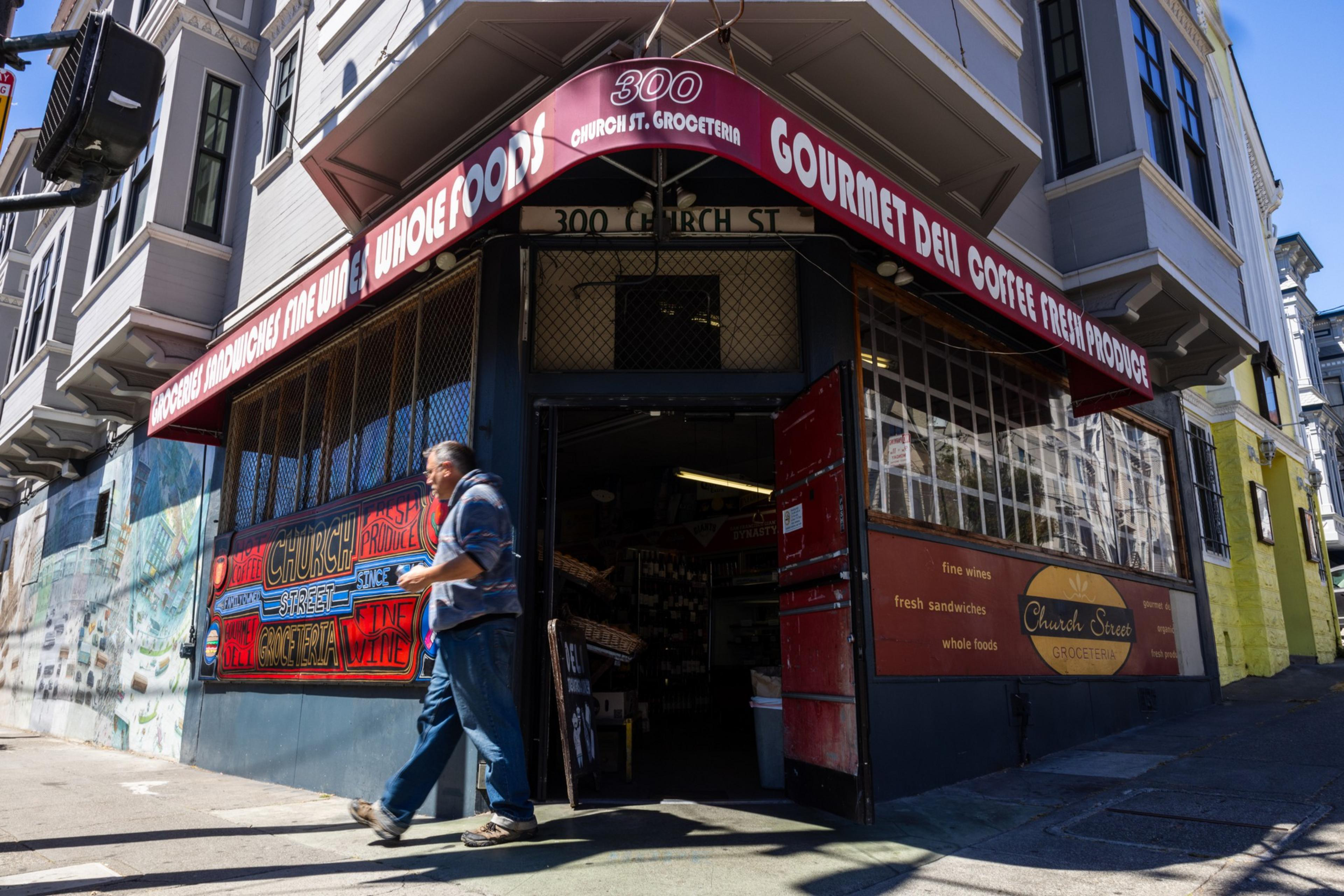 A person walks past a corner grocer with a red awning reading "300 Church St. Groceteria" and advertising gourmet deli, coffee, and fresh produce.