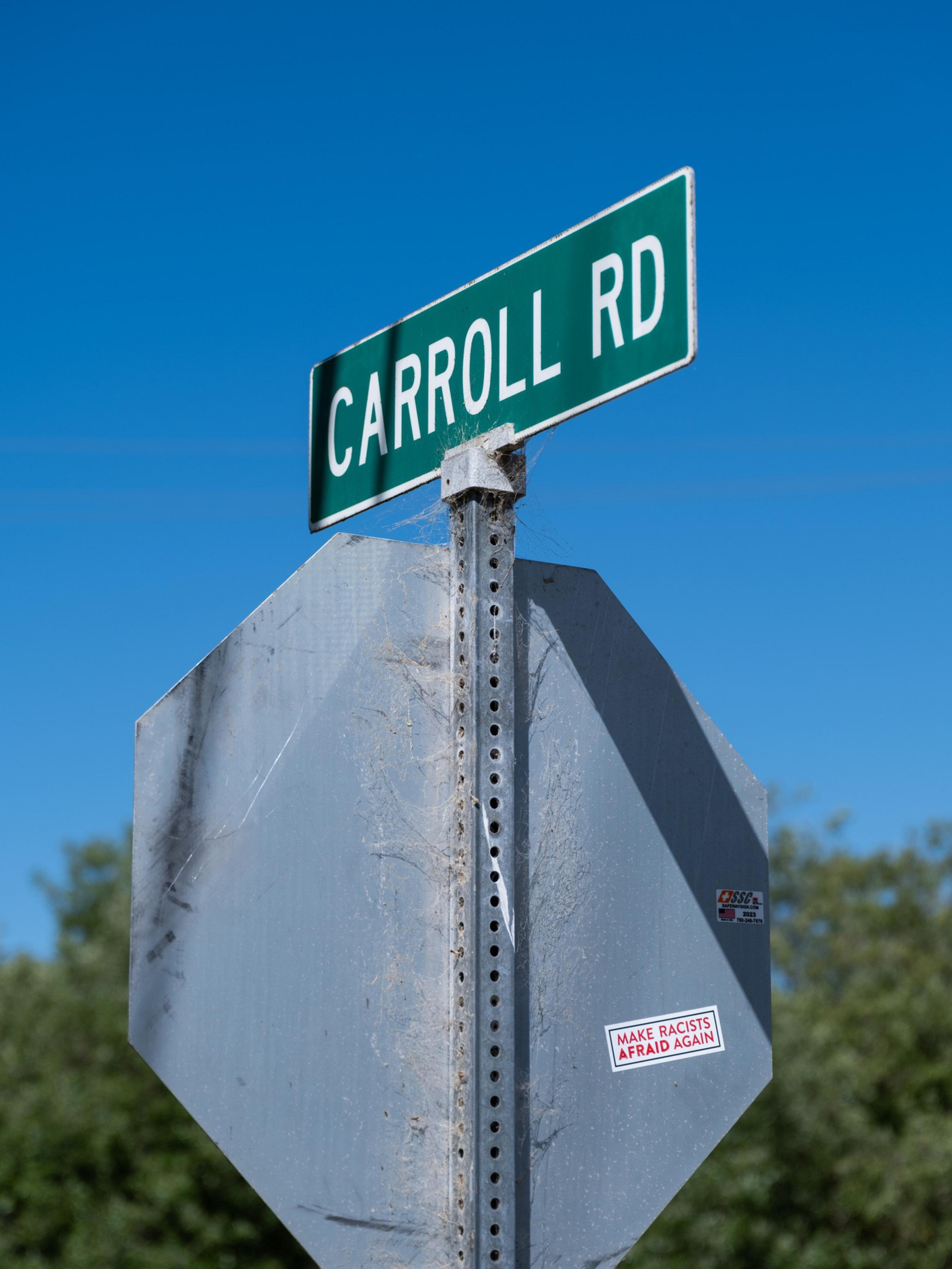 A street sign labeled &quot;CARROLL RD&quot; stands above a worn stop sign with a sticker that says &quot;MAKE RACISTS AFRAID AGAIN,&quot; set against a clear blue sky.