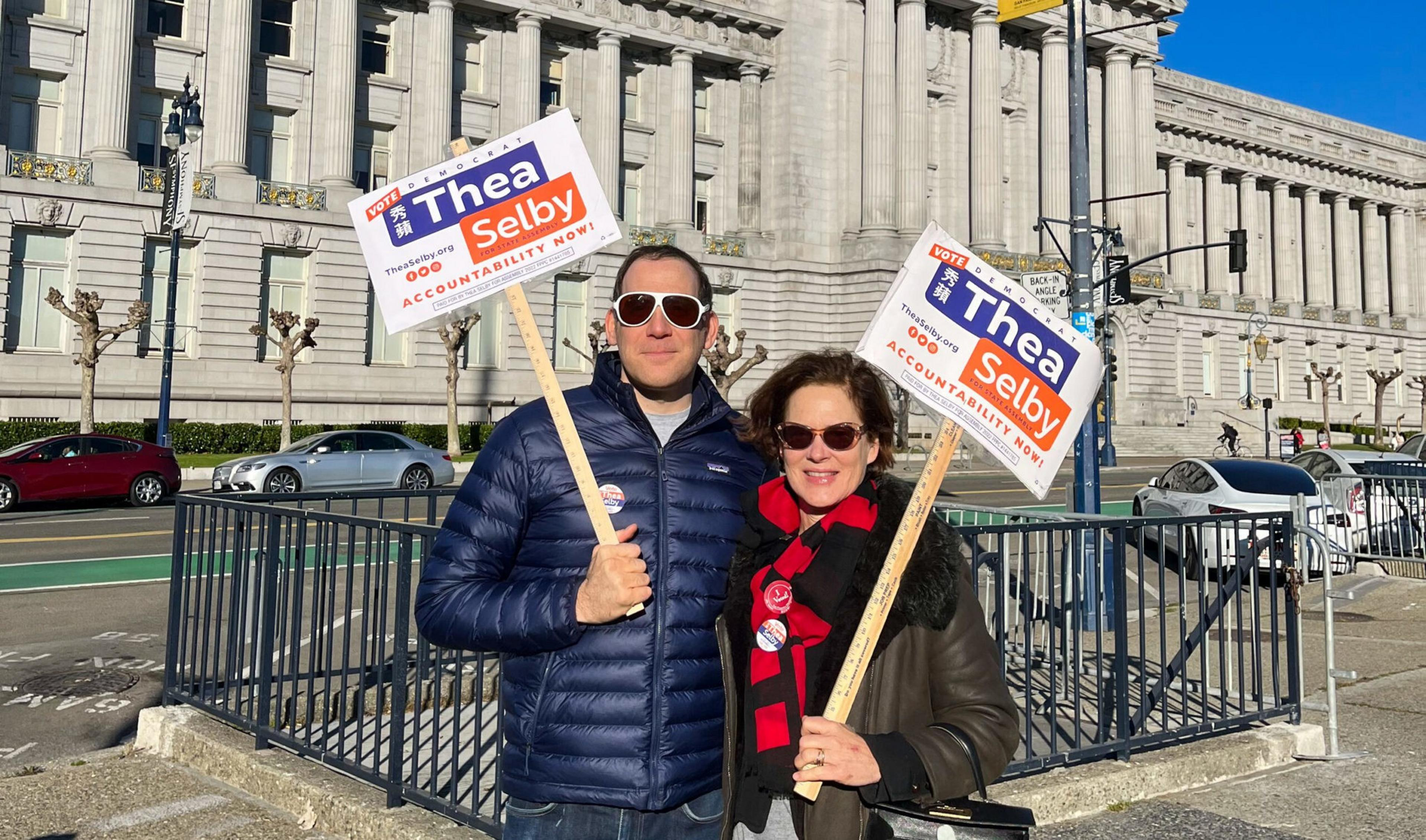 A man and woman wearing sunglasses hold signs that read "Vote Thea Selby Accountability Now!" outside a large, ornate government building.