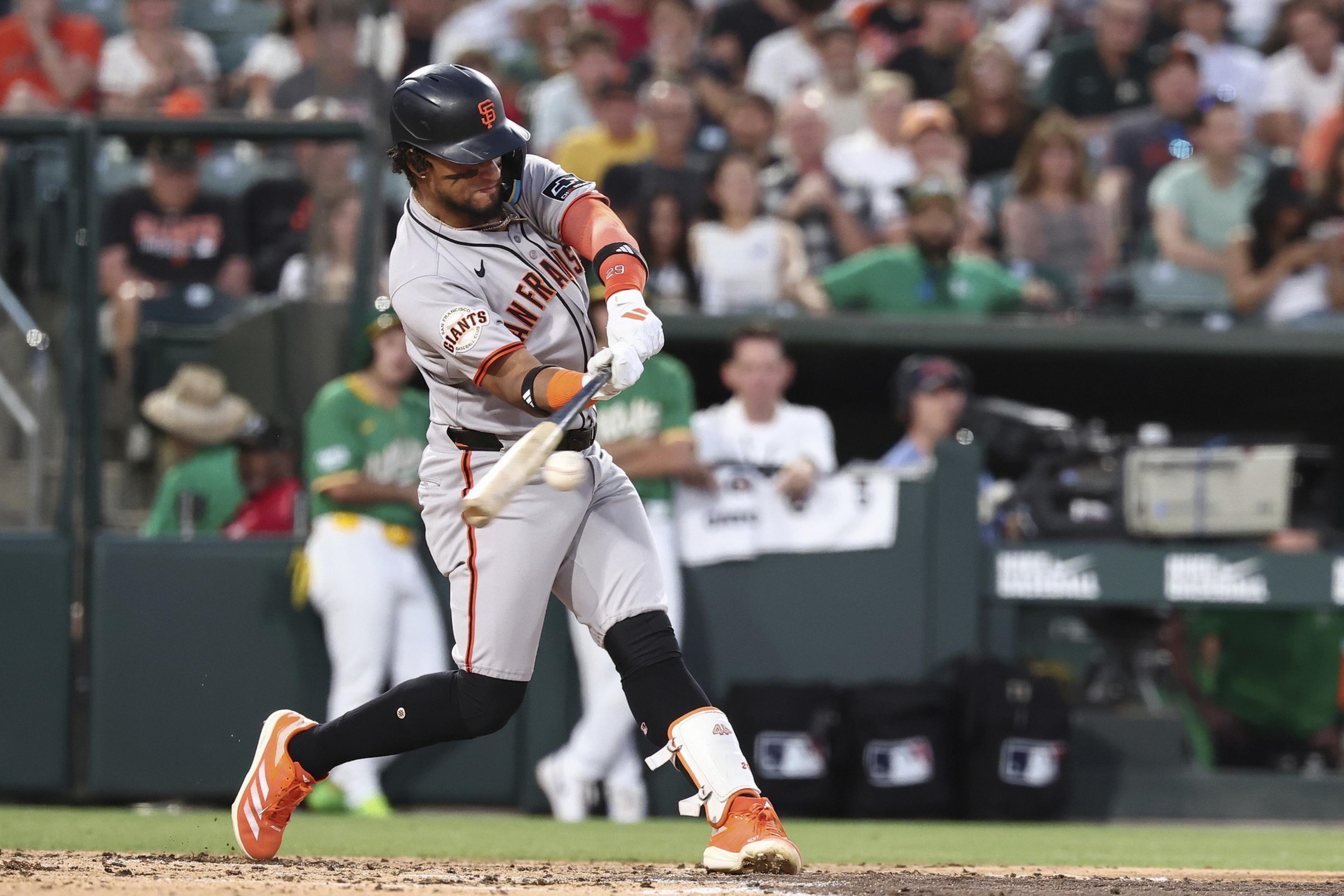 A baseball player in a gray jersey and orange helmet is swinging a bat, connecting with the ball. The background shows spectators and green-clad individuals.
