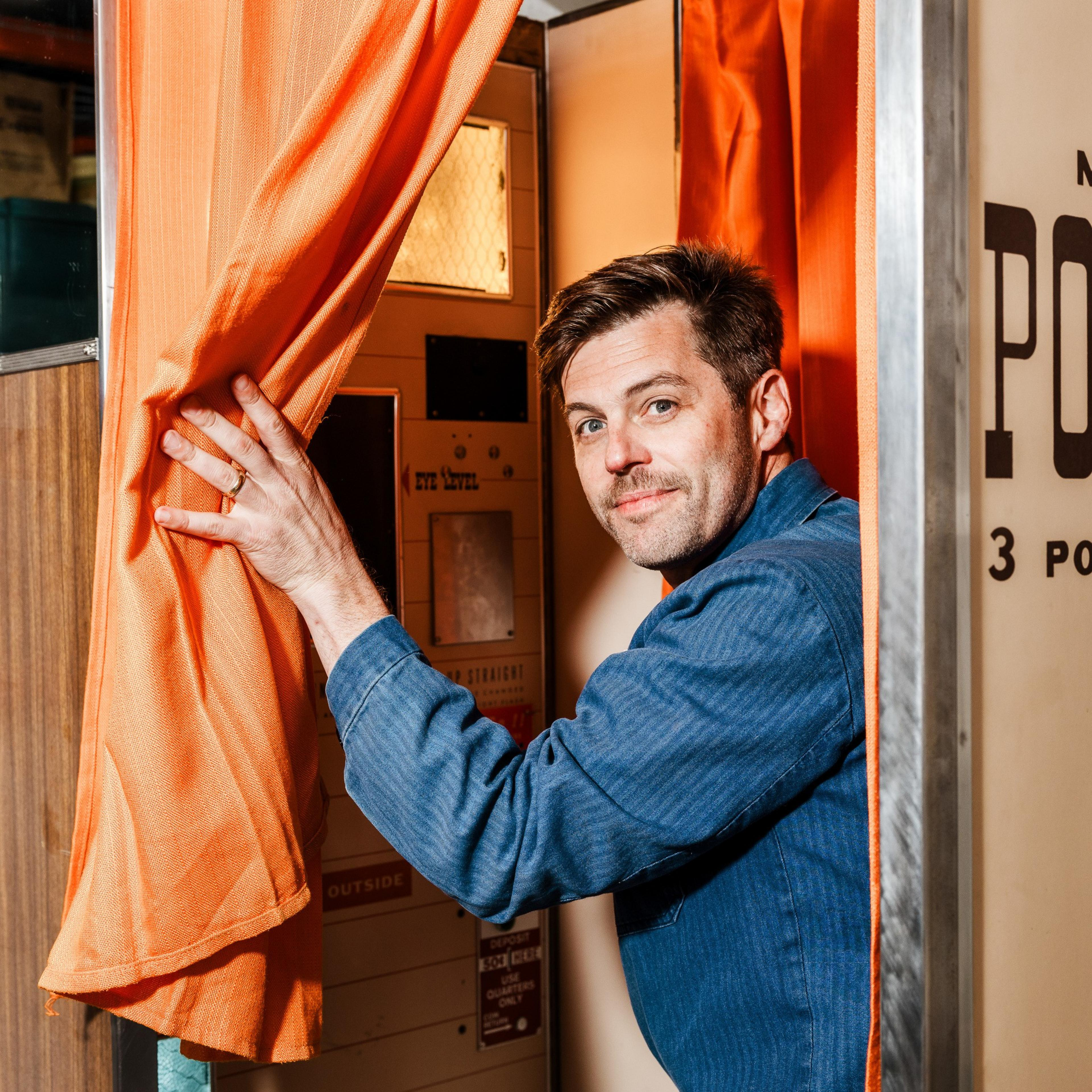 A man with short, dark hair and a light stubble peeks out from behind an orange curtain, holding it open with one hand. He stands inside a photo booth.