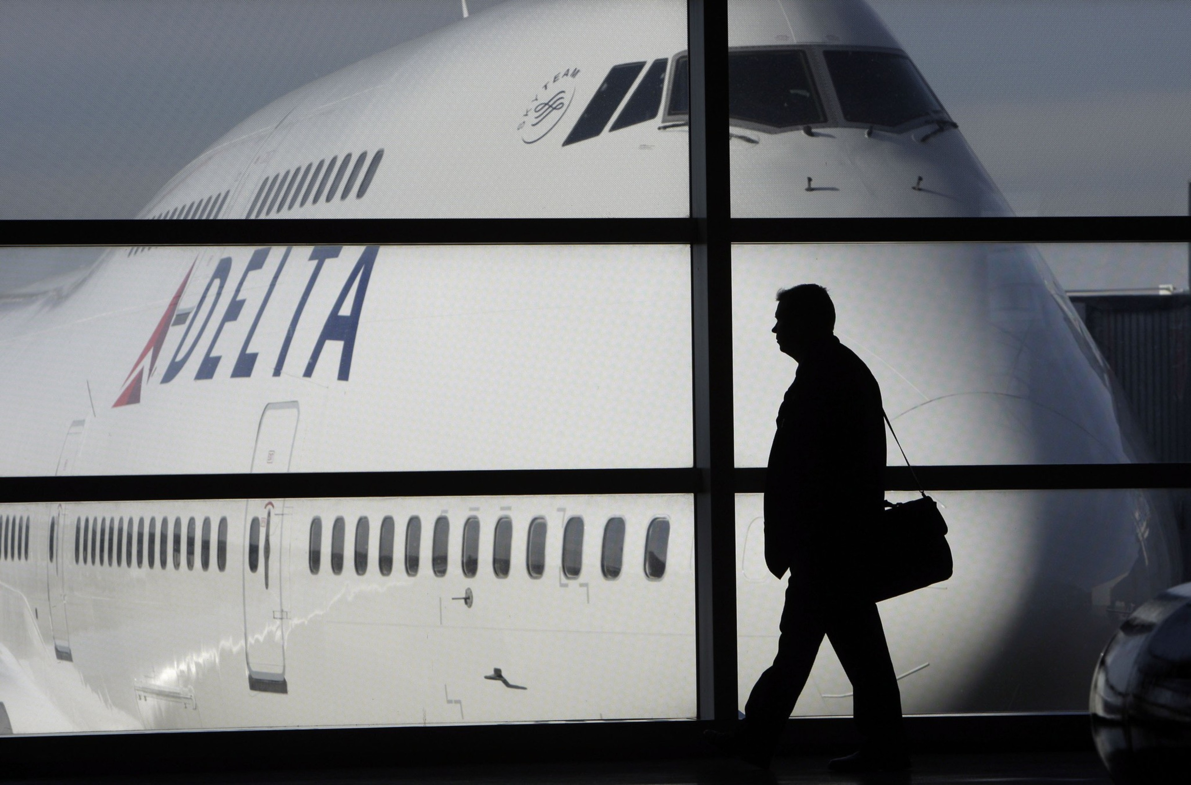 A large Delta airplane is parked outside a window, with the silhouette of a person carrying a bag walking inside the terminal.