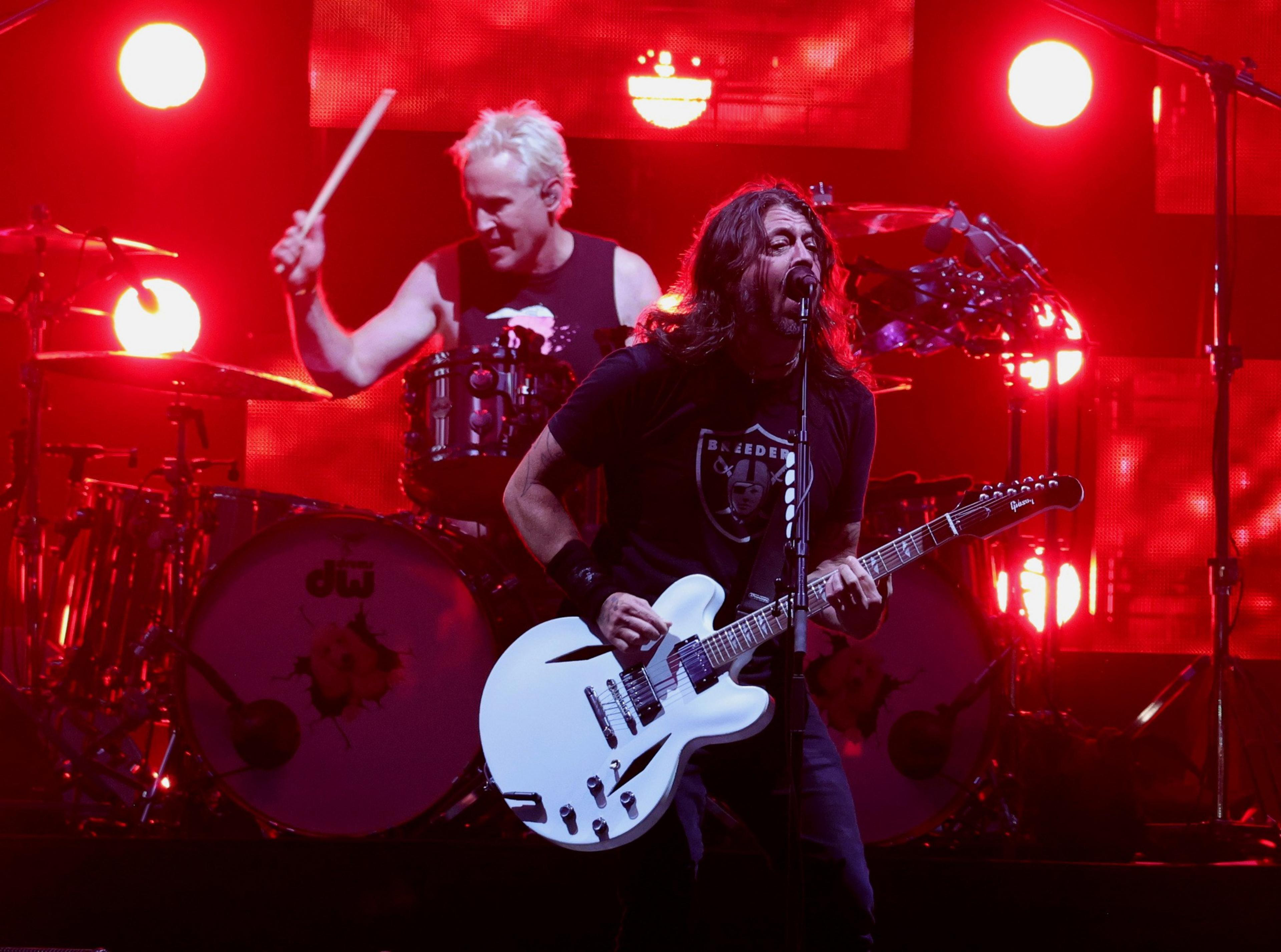 A guitarist passionately sings into a microphone while playing a white electric guitar as a drummer plays behind him with red stage lights glowing.