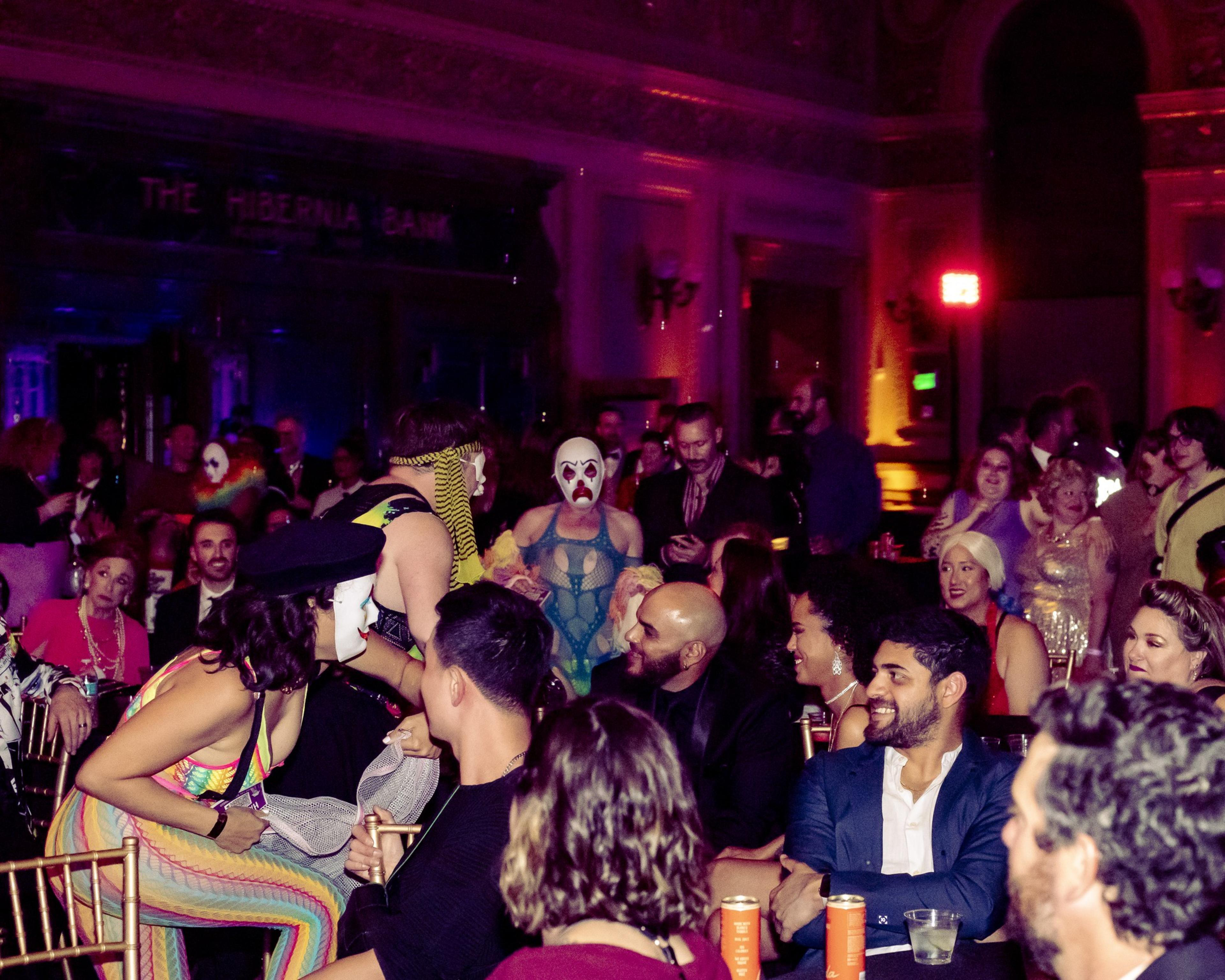 A lively crowd at a party with people in colorful outfits and face paint, seated and standing in a dimly lit room with ornate decor and an atmosphere of excitement.