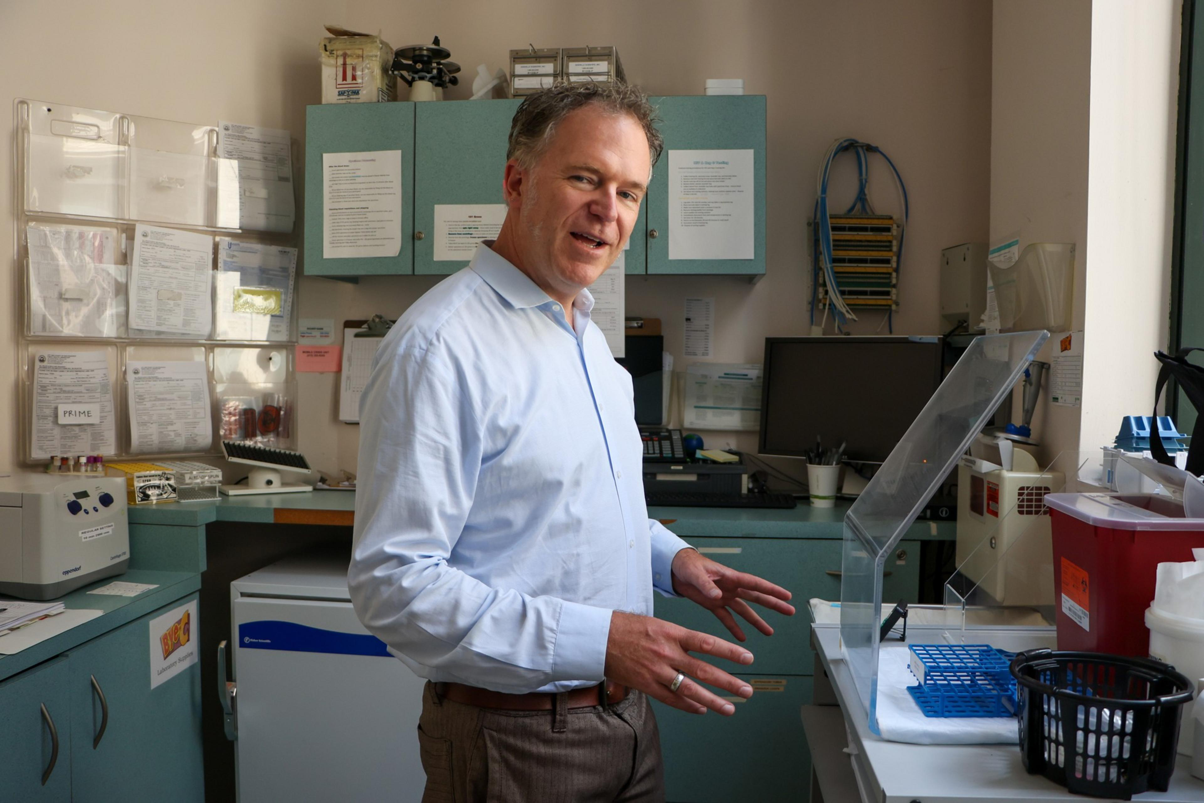 A man wearing a light blue shirt stands in a cluttered laboratory, with various equipment, documents on the wall, and cabinets and countertops around him.