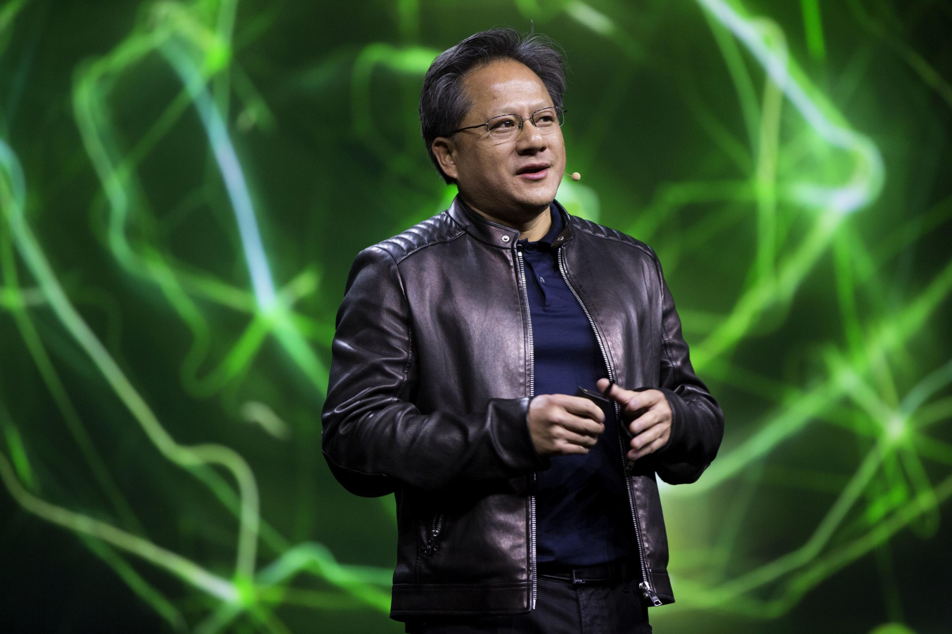 A person is speaking on stage with dynamic green lighting in the background. They're wearing glasses and a black leather jacket.
