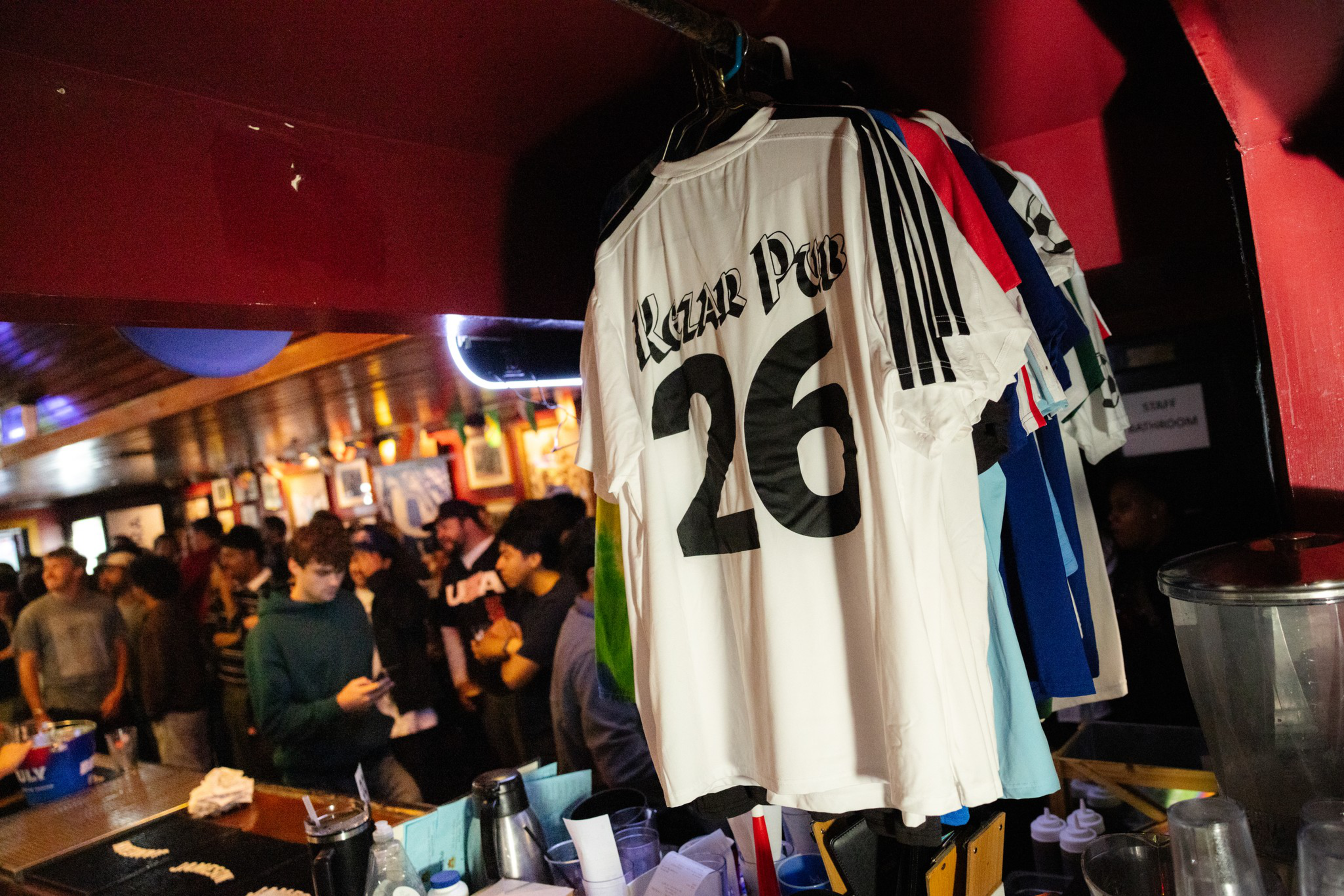 A white sports jersey with “Rozar Pub” and the number 26 hangs alongside other jerseys above a cluttered bar counter with people socializing in the background.