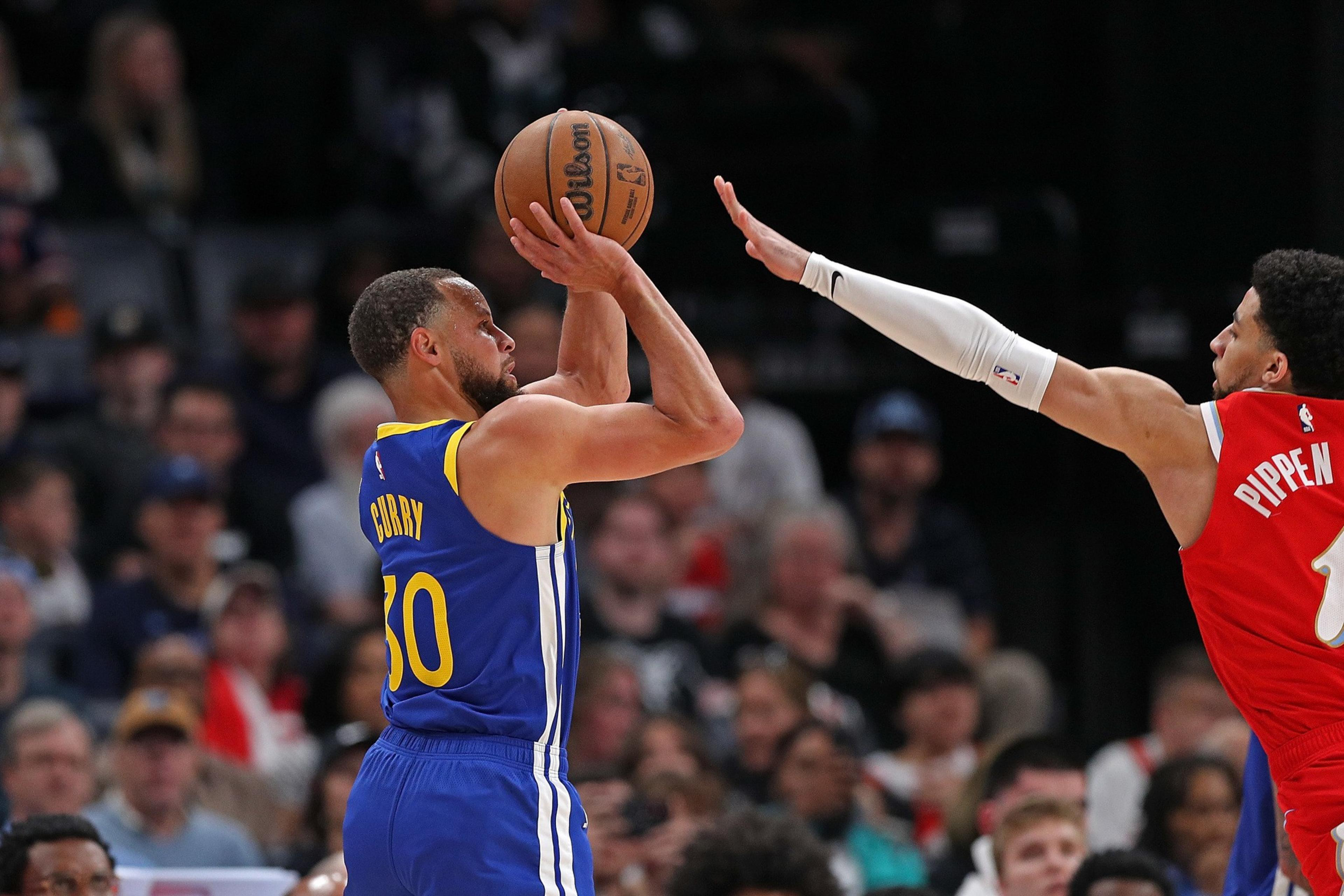 A basketball player in a blue jersey is shooting the ball while a player in a red jersey attempts to block the shot. The crowd watches in the background.