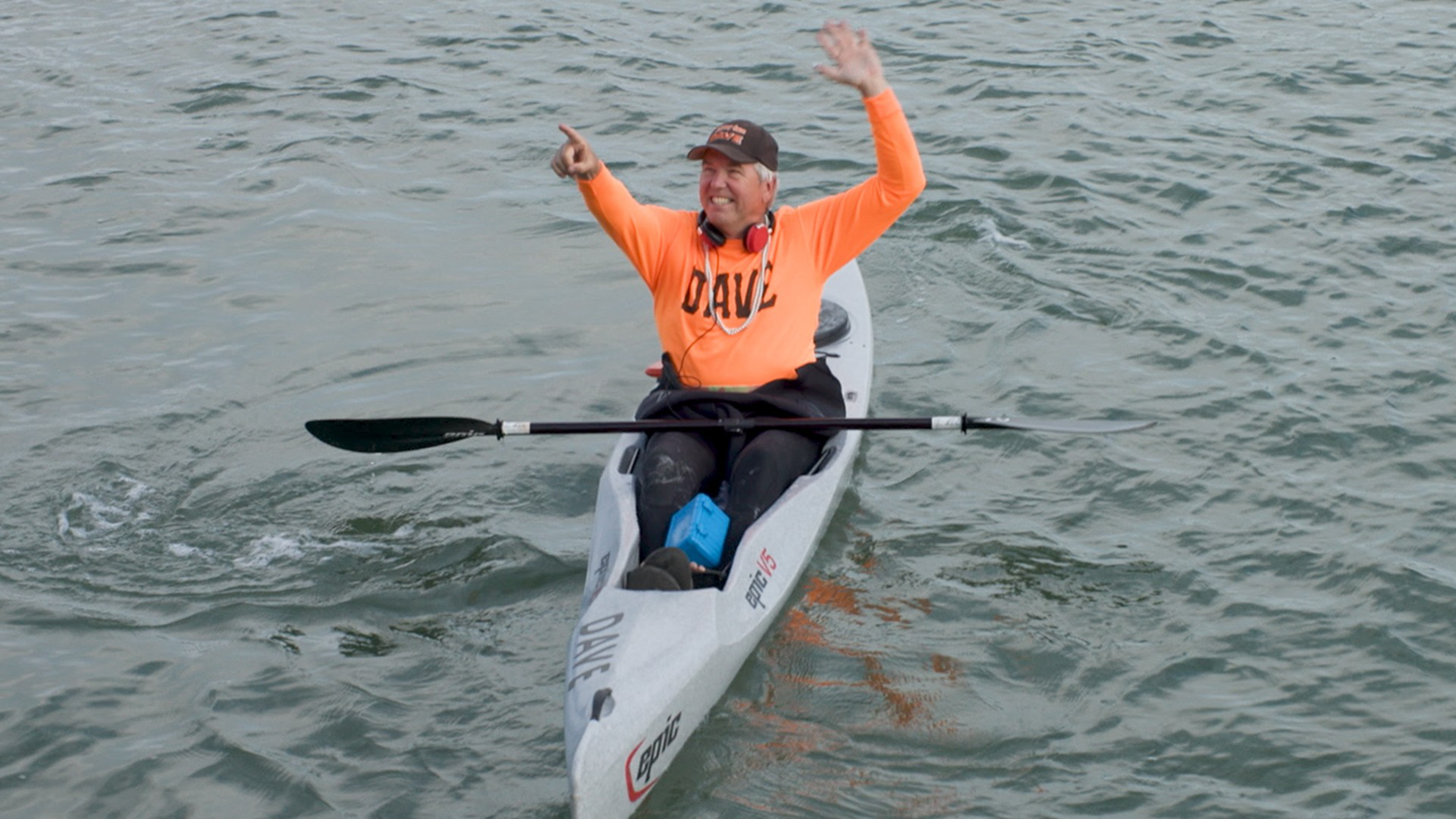 A person wearing an orange shirt and black cap sits in a kayak on the water, smiling and waving with both hands while the paddle rests across their lap.