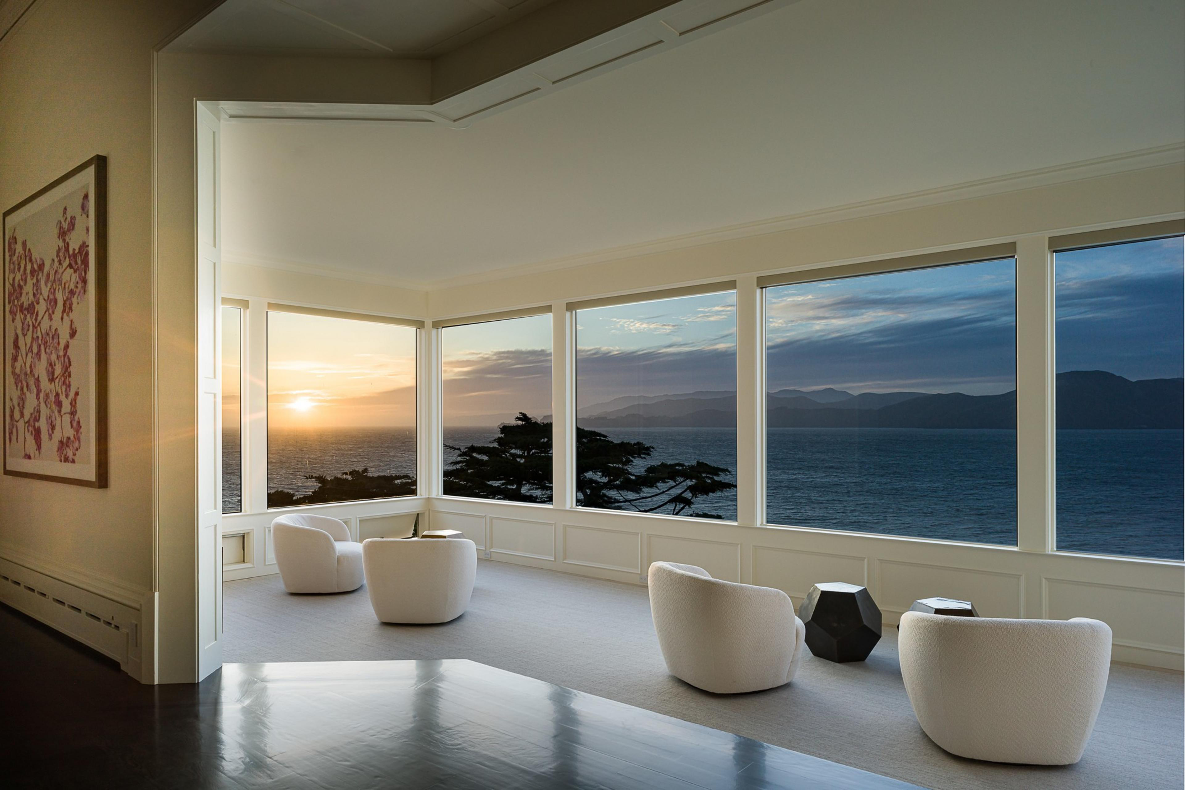 A serene room with large windows reveals a stunning ocean sunset view. Three white chairs face the view, and abstract art hangs on the wall.