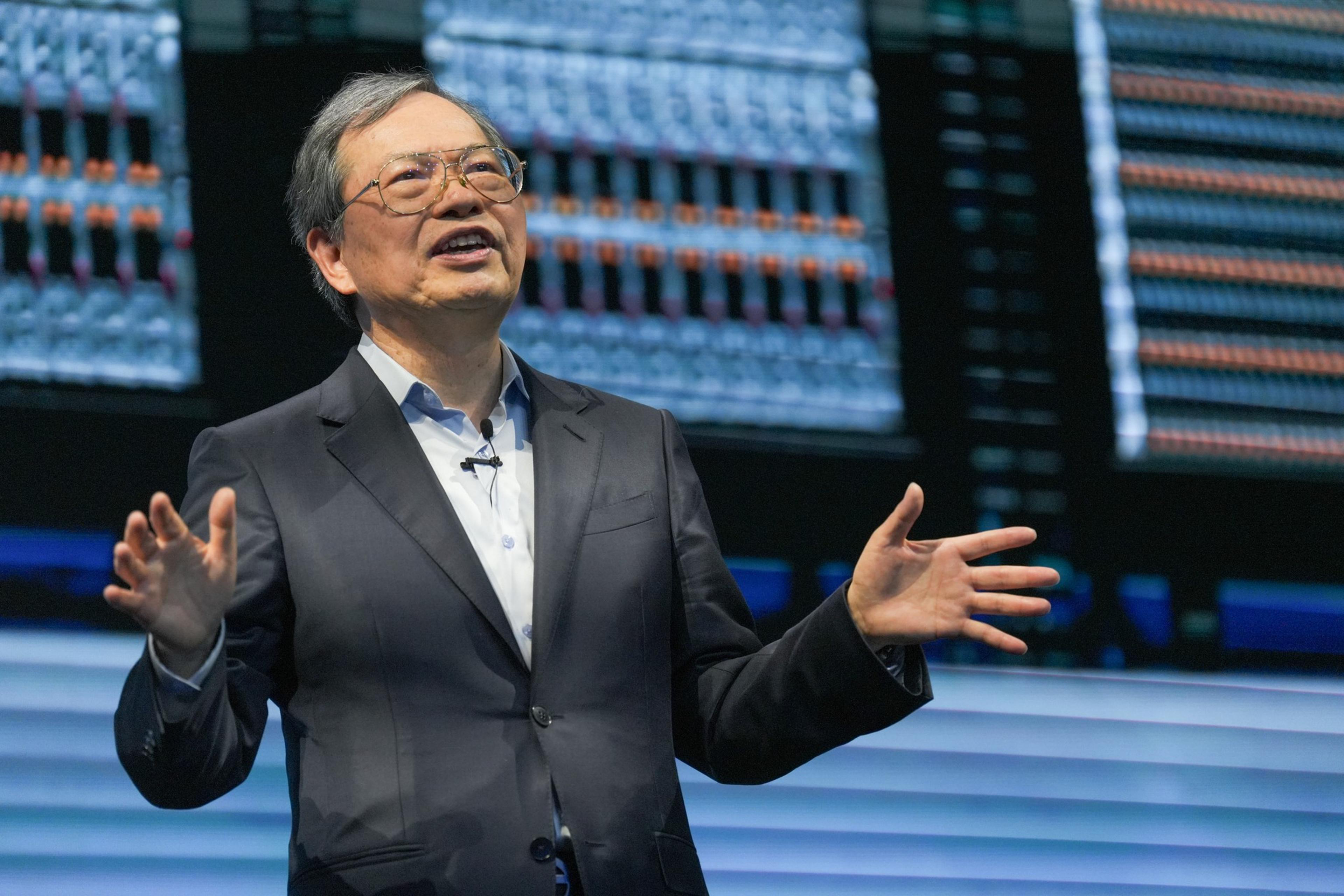 A man is on stage speaking, with large LED screens behind him, displaying colorful data racks.