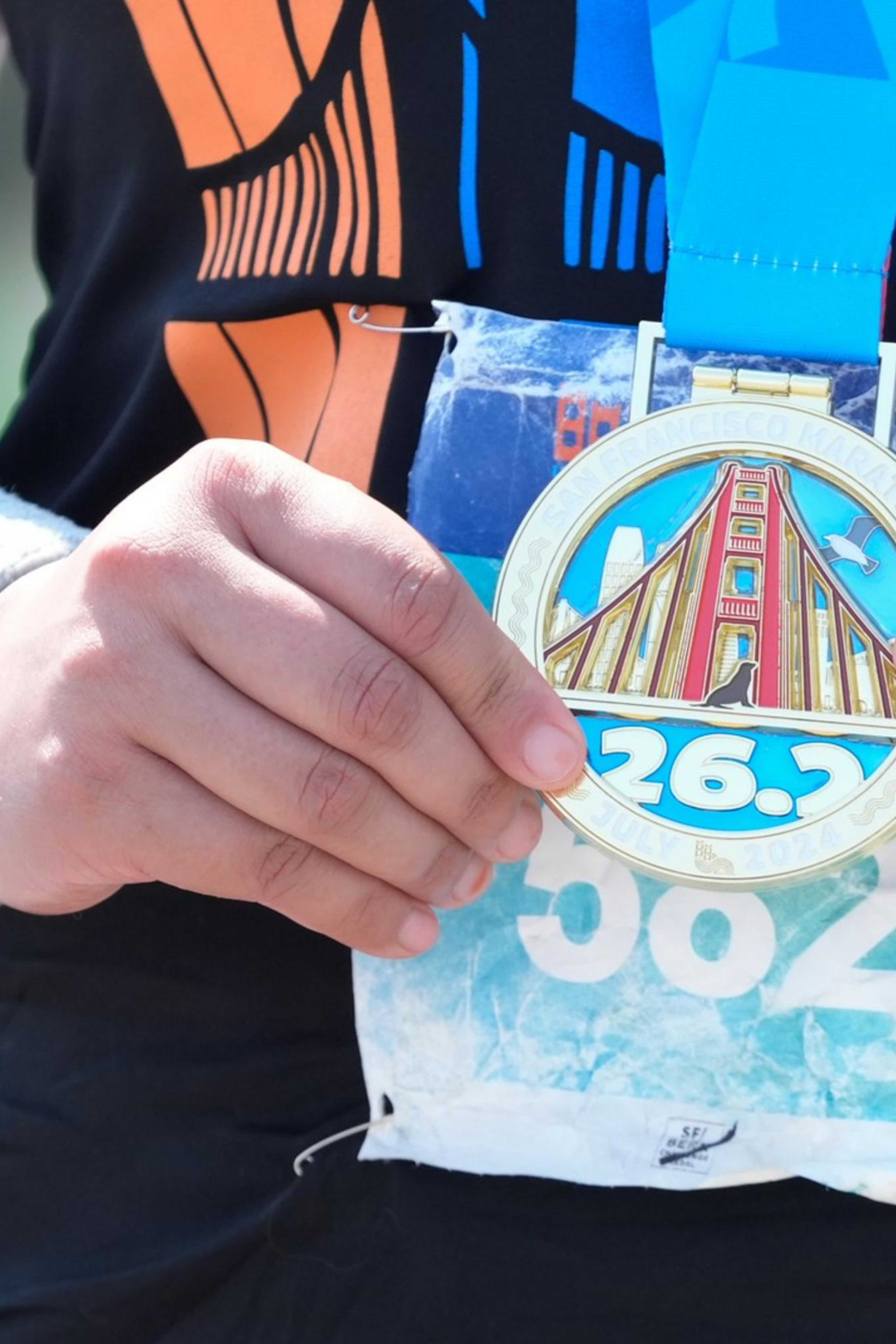 A person wearing a black shirt and a fitness tracker holds a marathon medal labeled &quot;26.2&quot; with an image of the Golden Gate Bridge and a bib number &quot;5820&quot;.