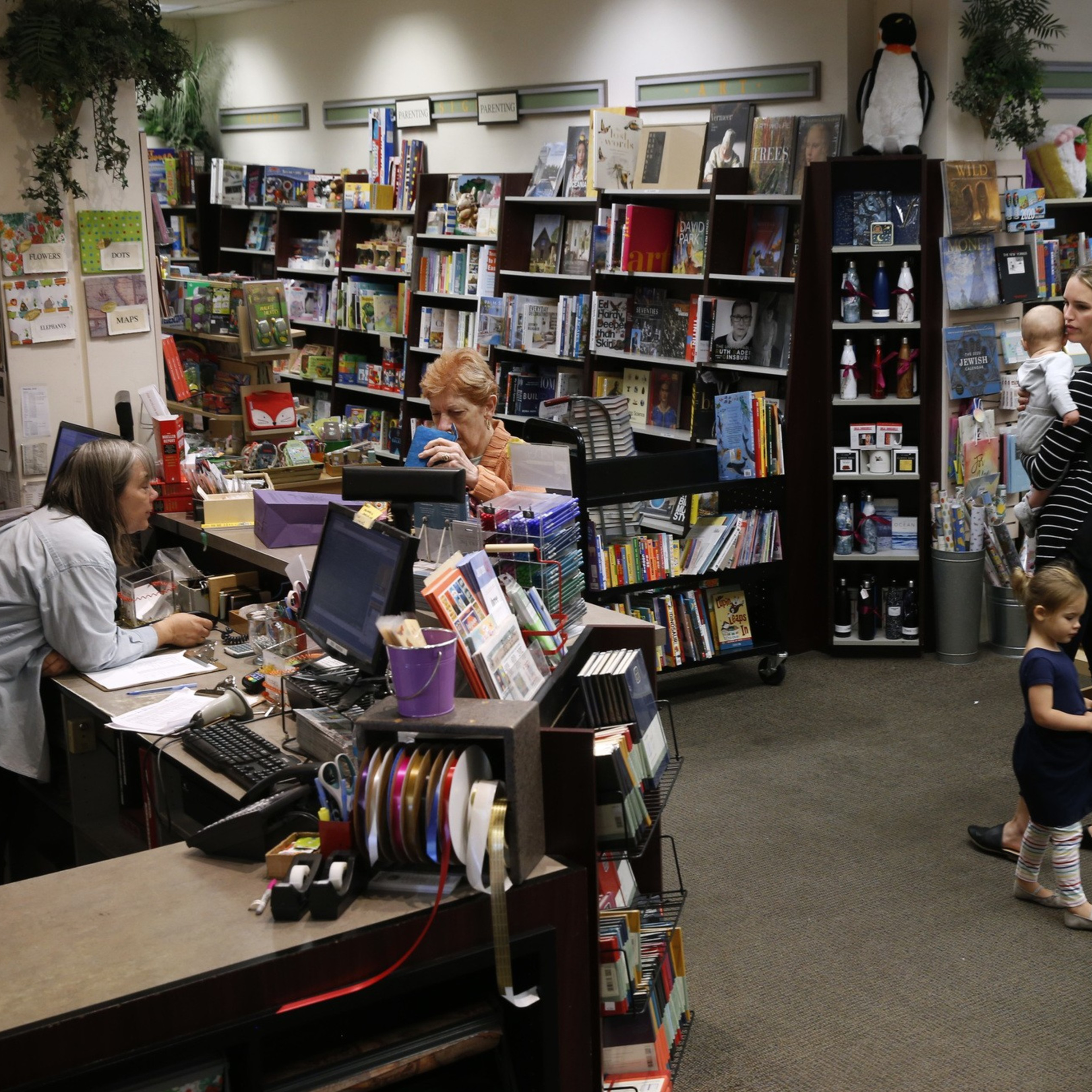 A busy bookstore with shelves full of books, two women at the counter, and a mother with two young children browsing nearby.