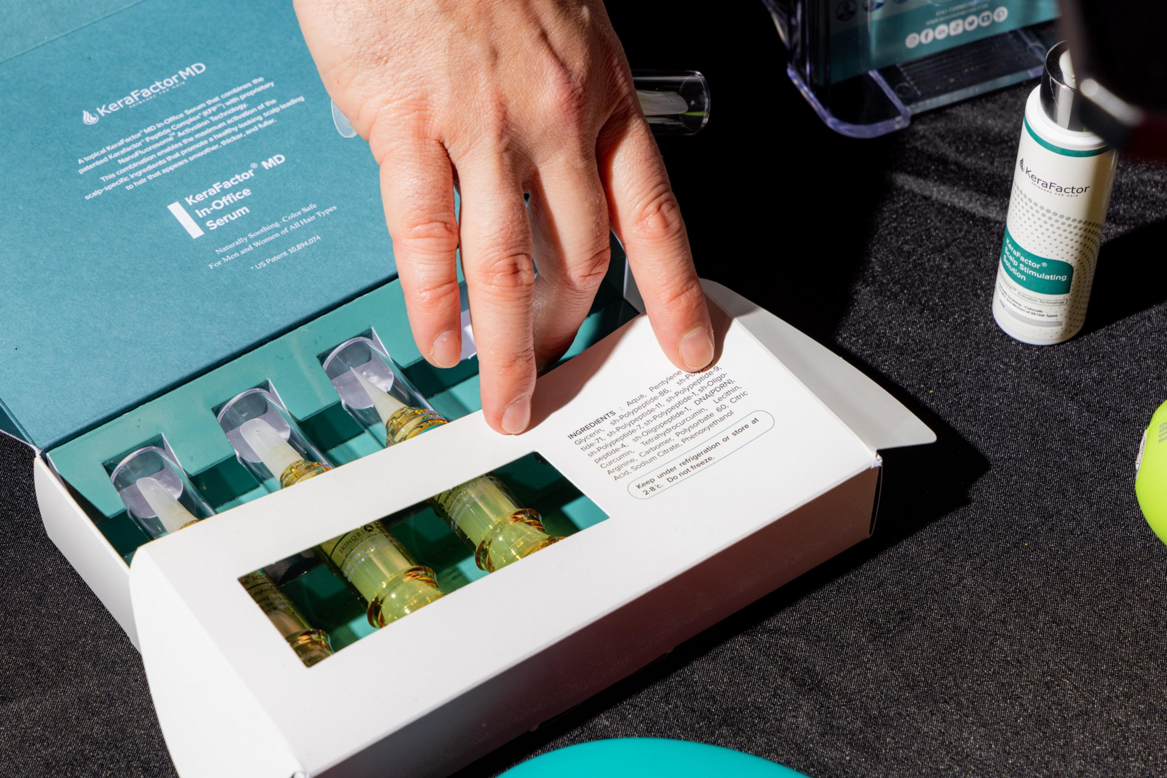 A hand is about to open a box labeled &quot;KeraFactor MD,&quot; containing four small bottles with droppers, against a dark surface, with another small bottle nearby.