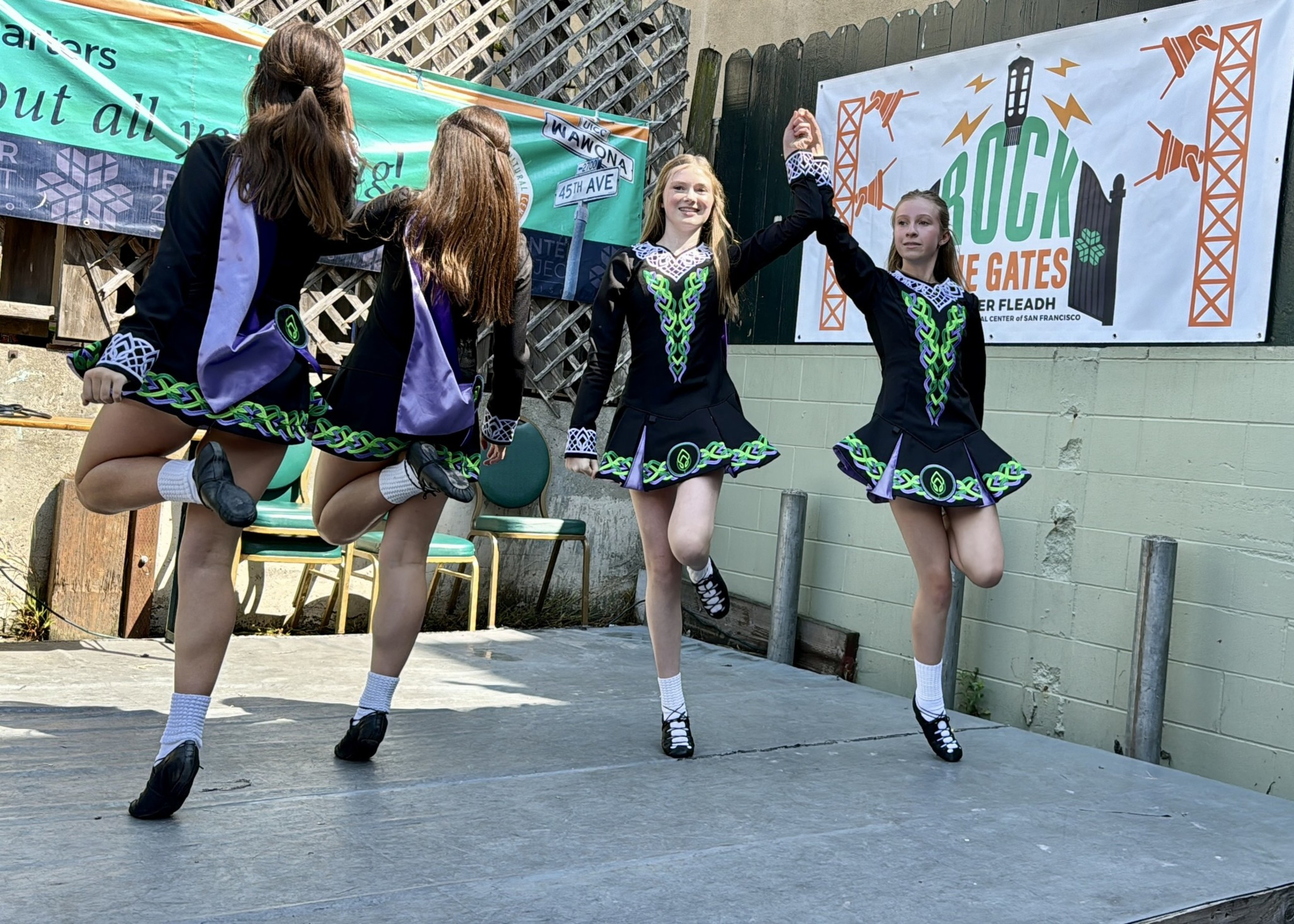 Four girls in black and green Irish dance dresses perform a step dance on an outdoor stage, two holding hands in the air.