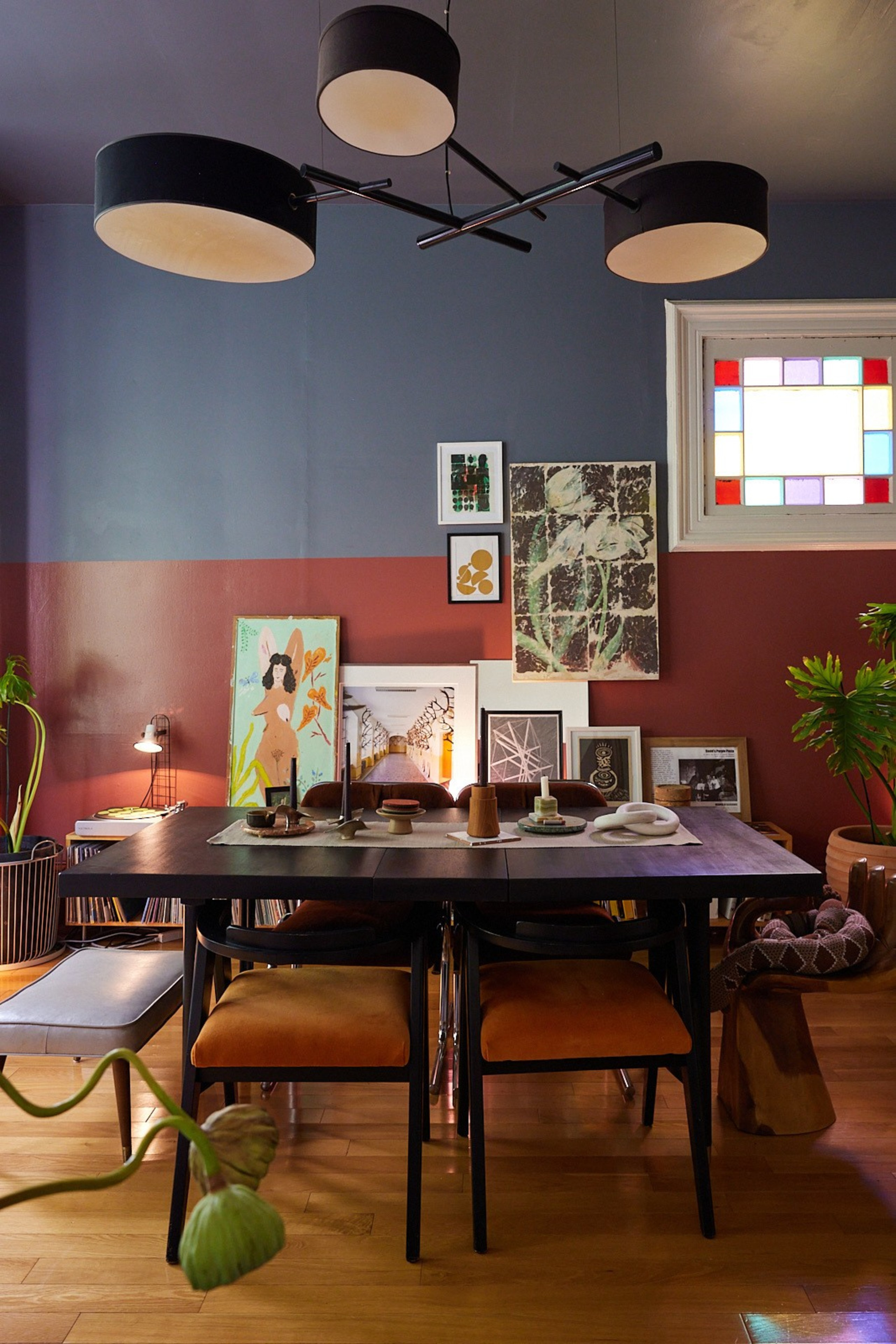 A dining room with a black table and orange chairs, adorned with various artworks on a blue and maroon wall. A modern chandelier hangs above, under a stained-glass window.