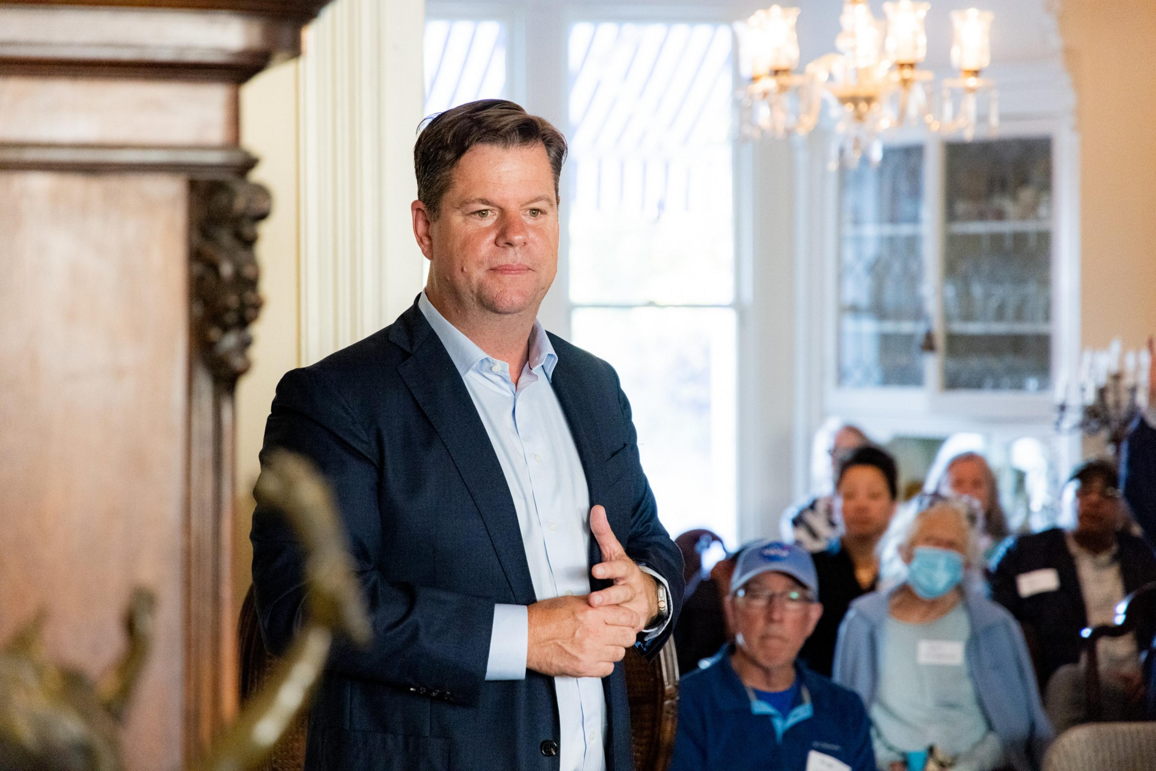 A man in a dark suit and light blue shirt stands in a room with a chandelier. Several seated people, some wearing masks and hats, are attentively watching him.