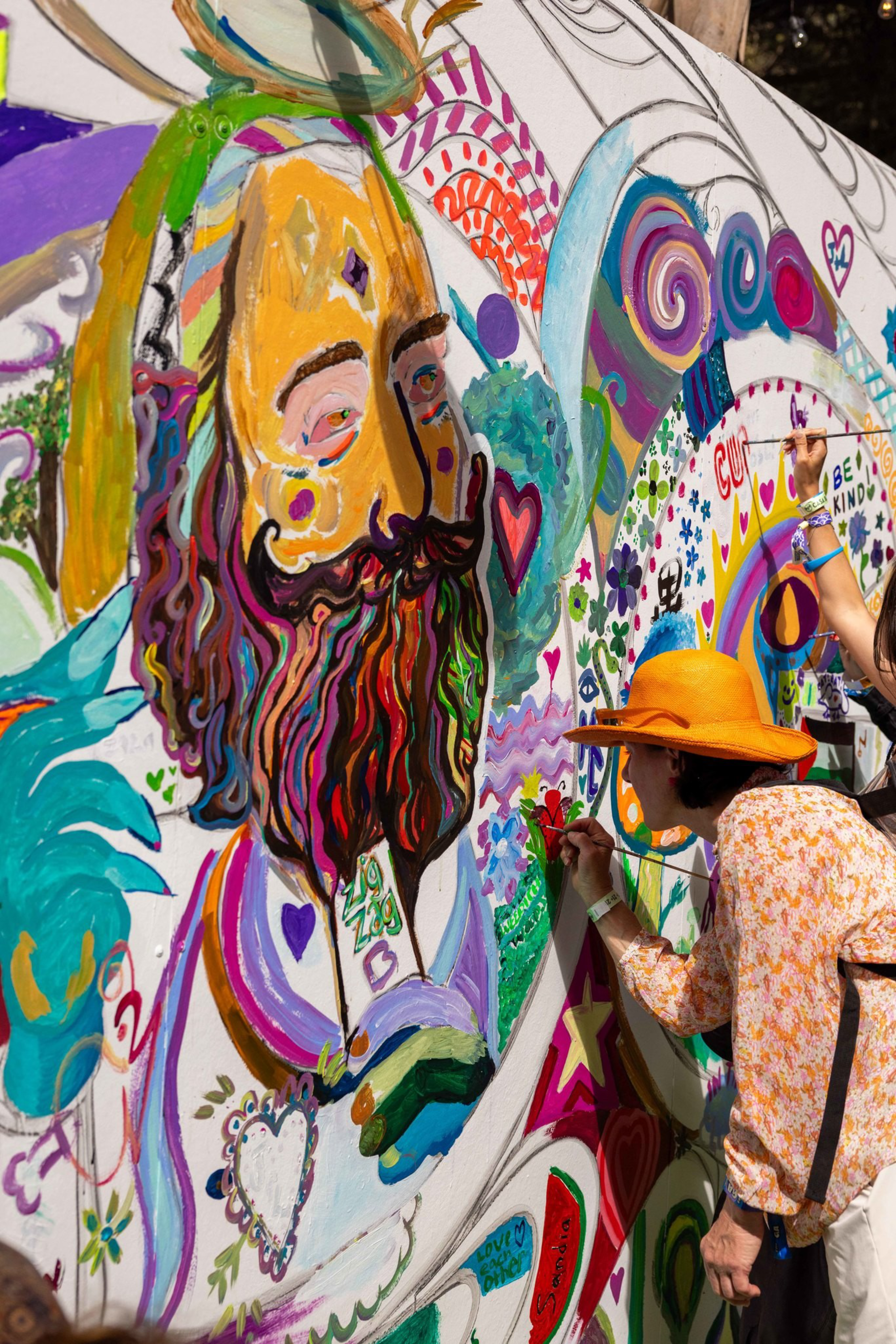 A colorful mural features a bearded man with vibrant patterns and hearts, while a person wearing an orange hat paints additional details on the wall.