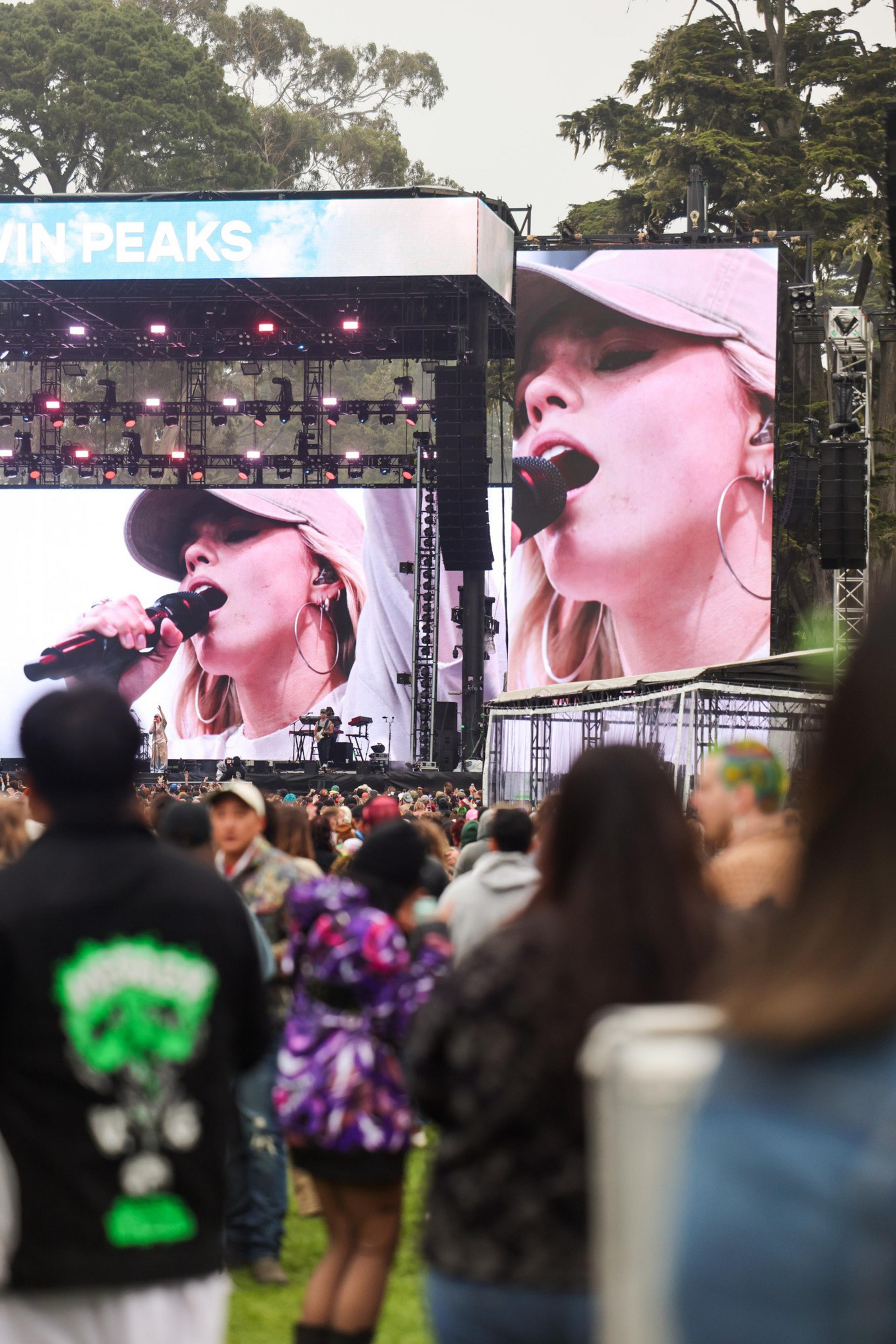 A large outdoor concert stage features a close-up of a female singer with a cap on a big screen, while a crowd of people watches. Trees are visible in the background.
