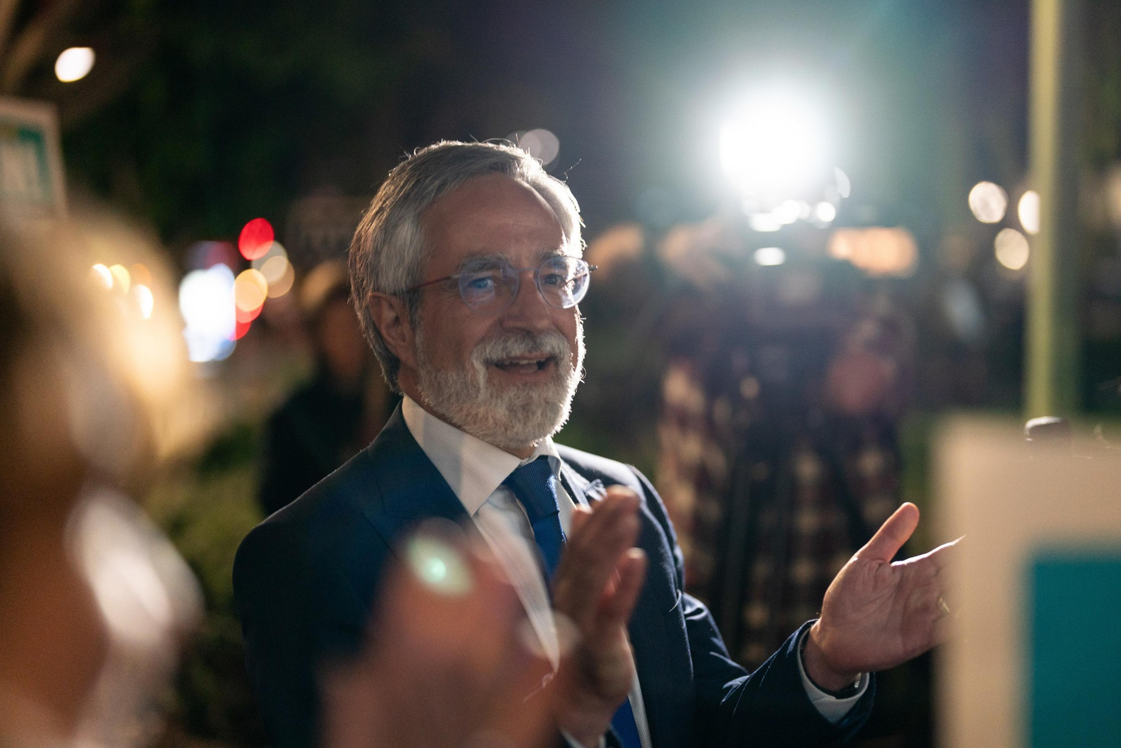 A man with glasses and a beard is smiling and clapping, illuminated by a bright light in the background. It appears to be night with blurred lights around.