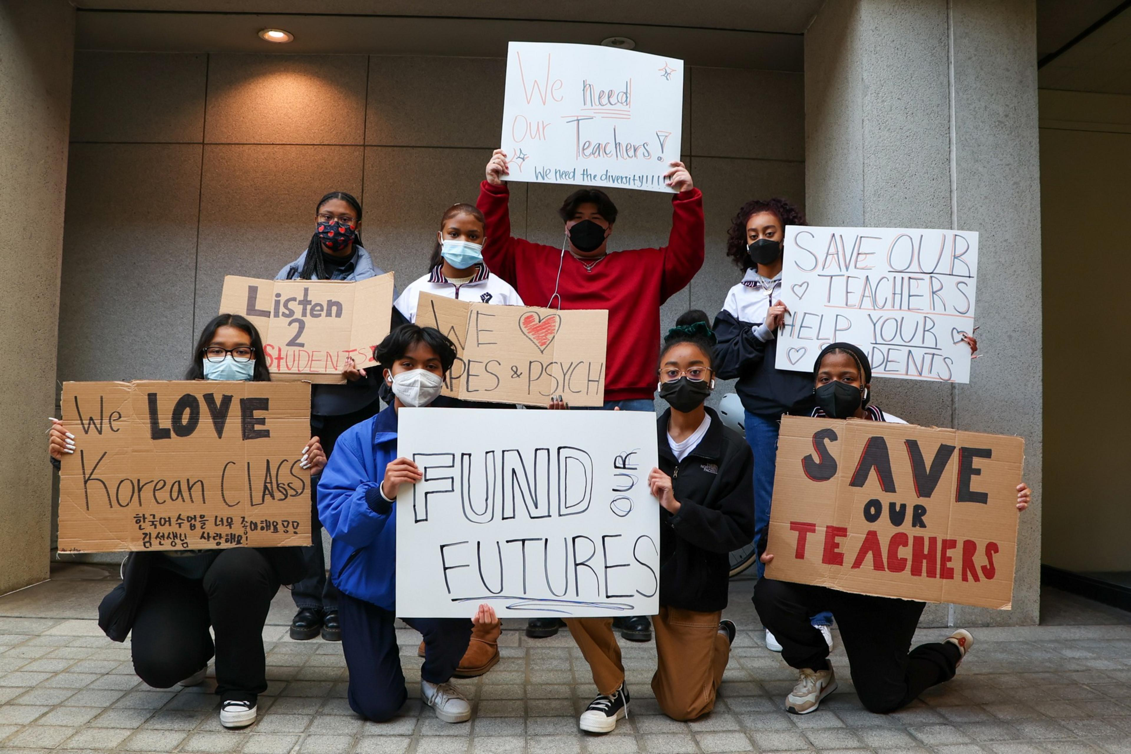 Eight students wearing masks hold signs with messages like "We love Korean class," "Fund our futures," and "Save our teachers" in a school setting.