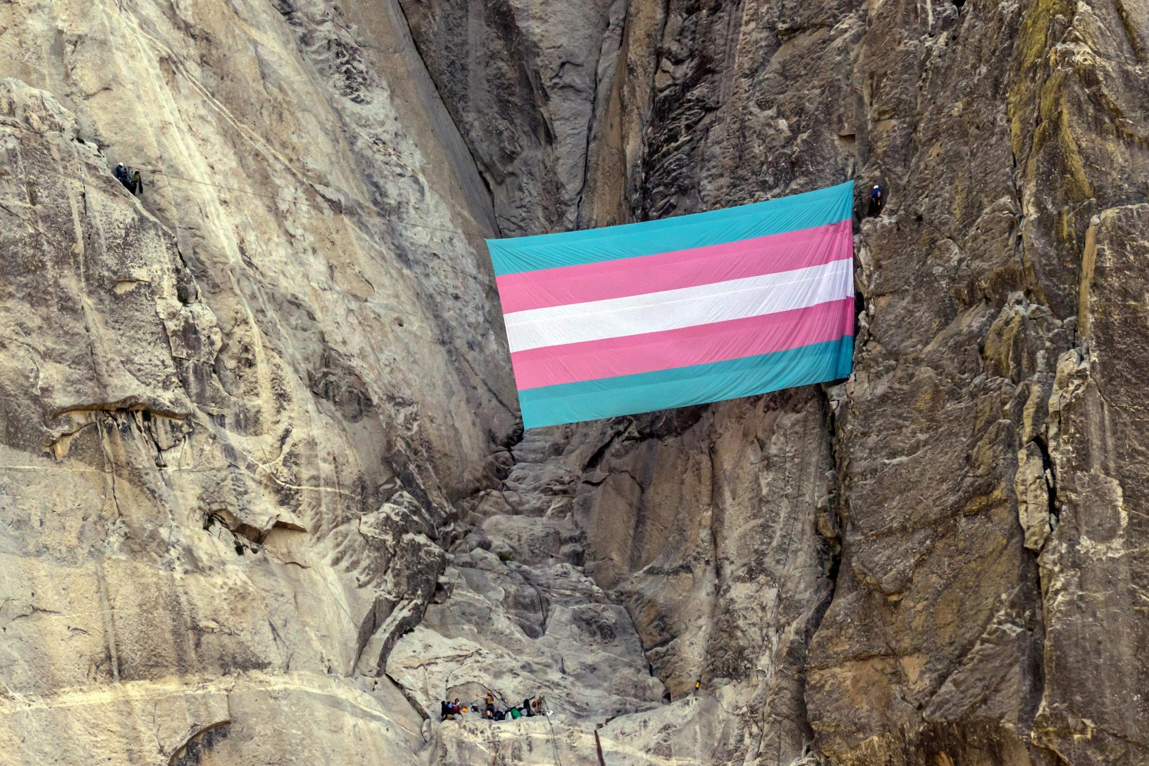 A large transgender pride flag hangs vertically between two steep rocky cliffs, held up by climbers visible on both sides.