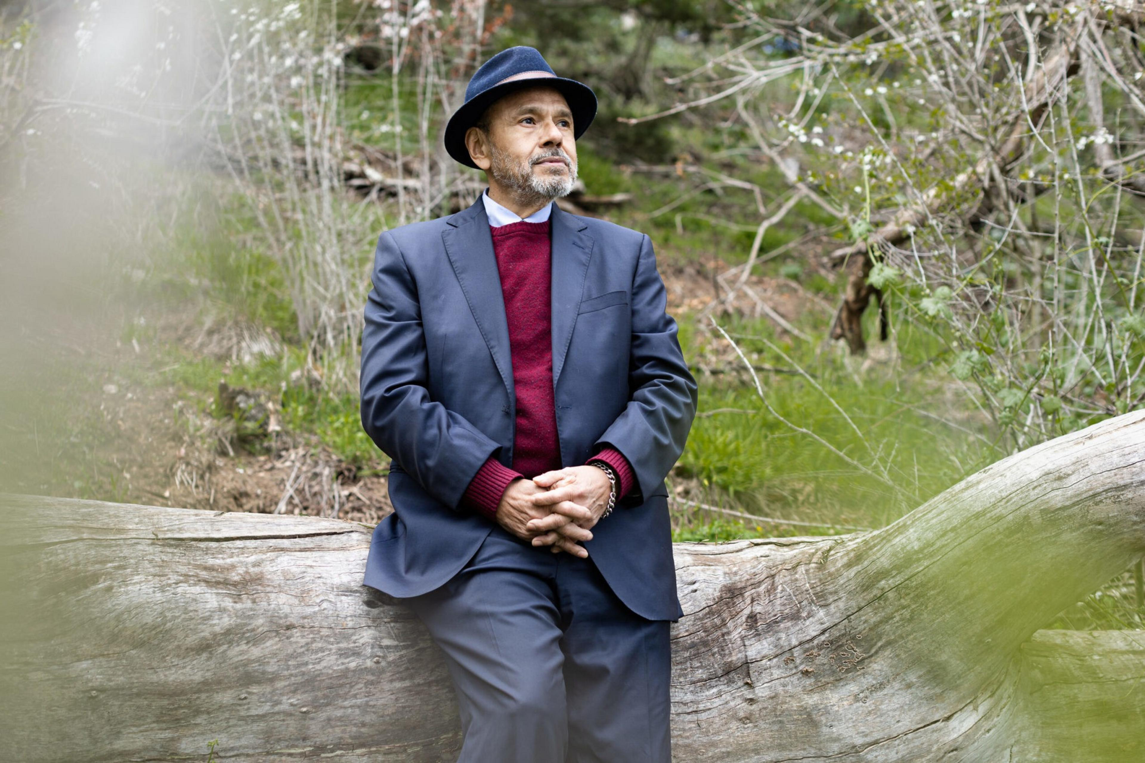 An elderly man wearing a black hat, navy blazer, and red sweater sits on a large fallen log in a quiet forest setting.
