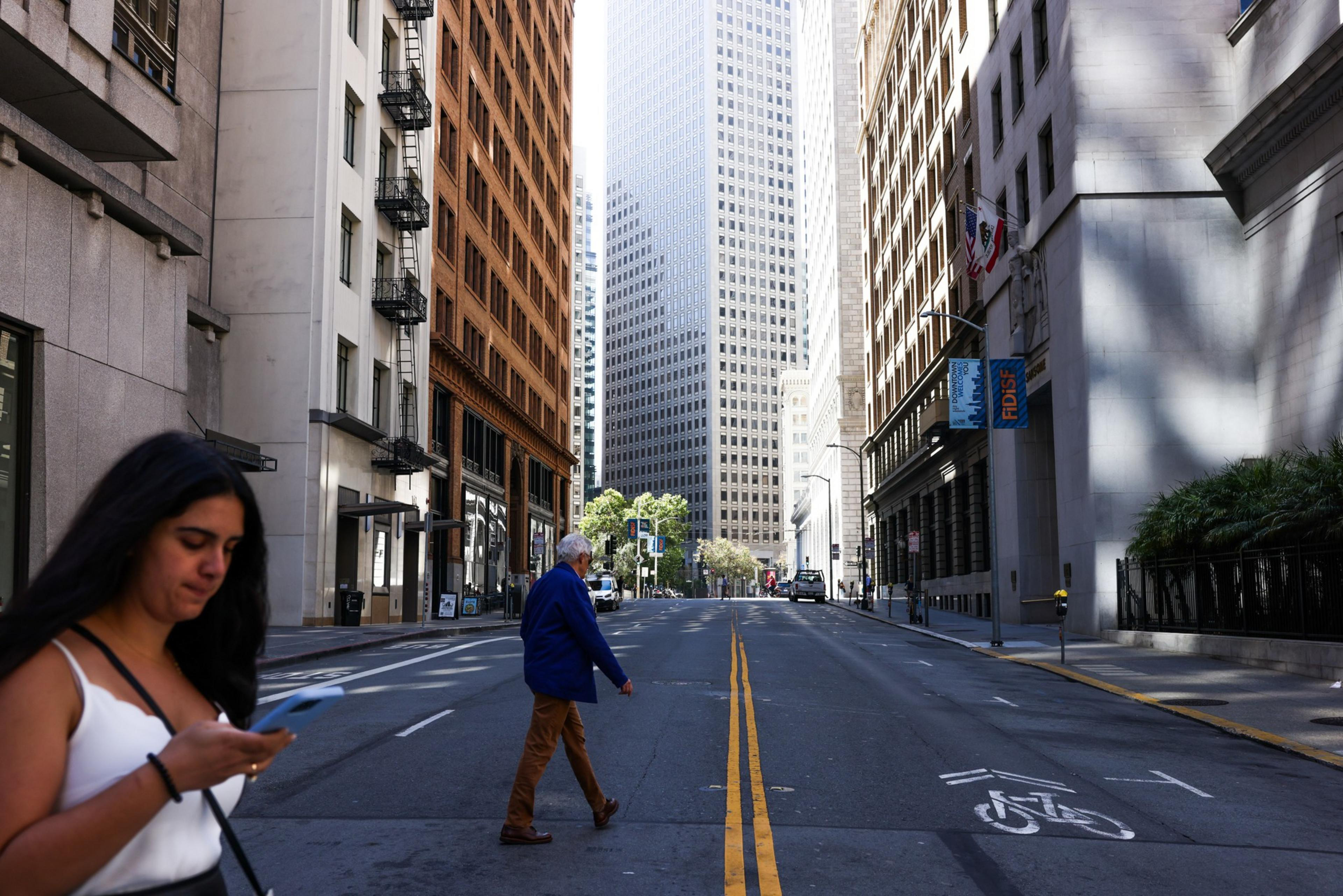 A woman looks at her phone on the left while an older man crosses an empty city street lined with tall buildings on both sides.